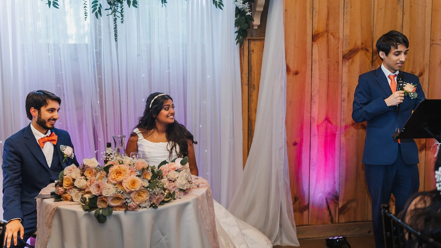 Natalia and Ansel smiling at their sweetheart table during a toast in the barn at Wolf Oak Acres