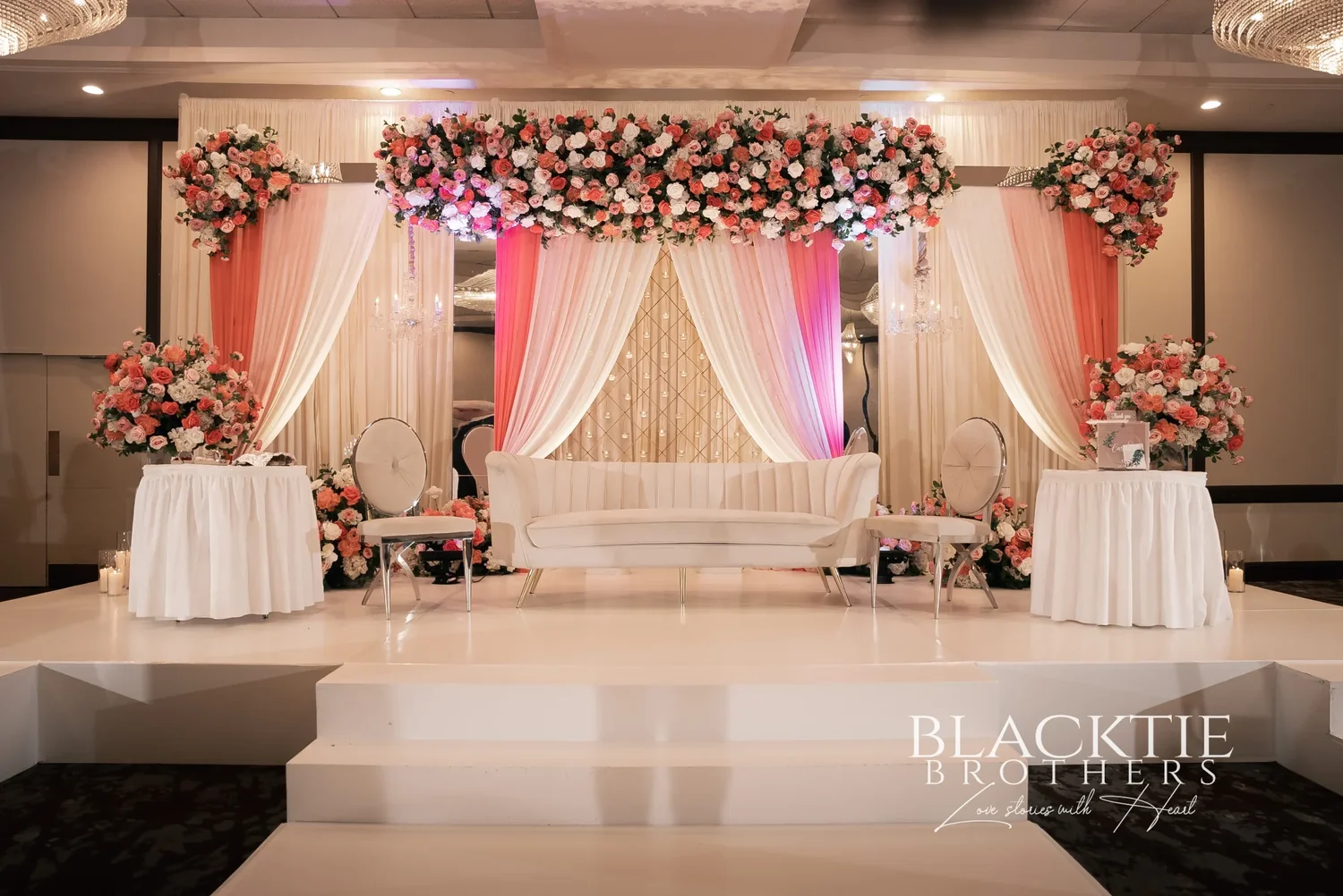 An ivory and pink draped wedding stage with flower arrangements, a white sofa and two chairs on a raised white platform in a ballroom at the DoubleTree by Hilton Binghamton