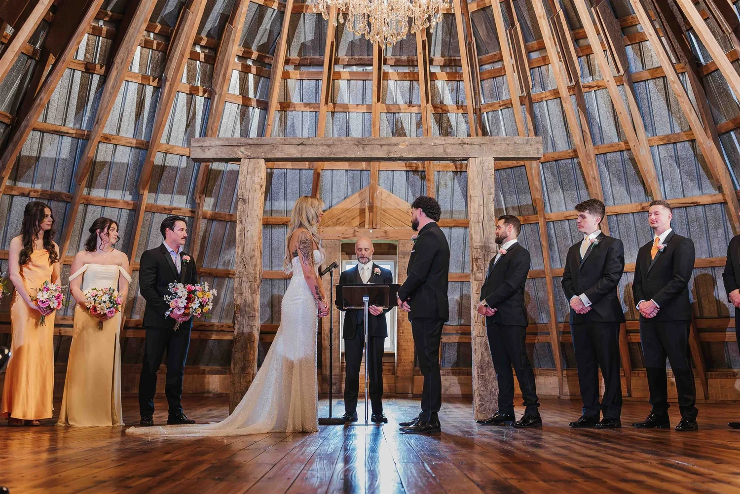 Kari and Josh face each other with their officiant and wedding party beneath the timber-framed ceremony roof.