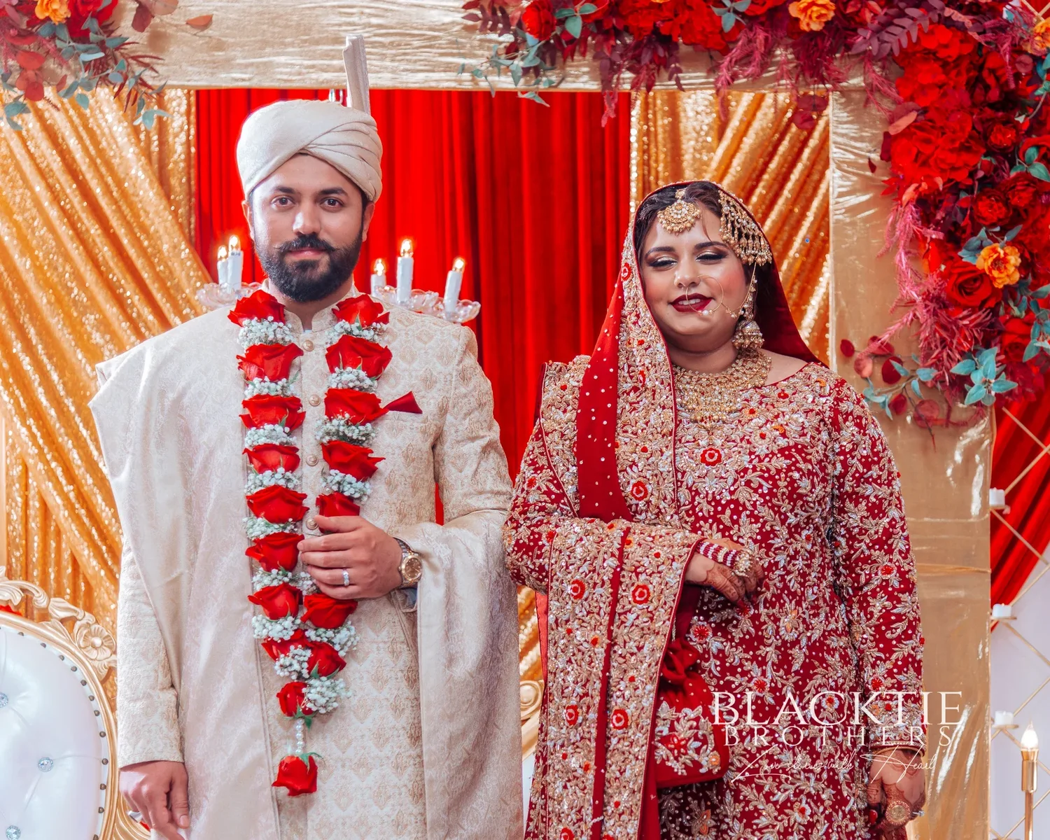 The bride and groom standing together on a red and gold draped stage, he in an ivory sherwani with a garland of red roses, she in red and gold