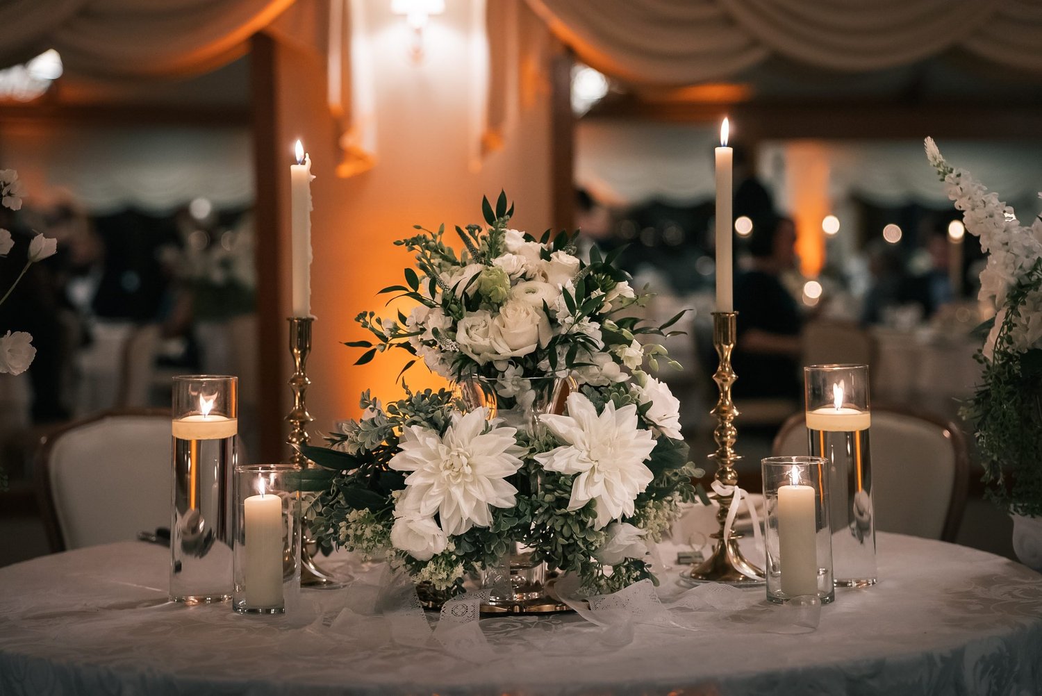 A reception table set with white flowers, tall gold candlesticks and candles glowing in glass
