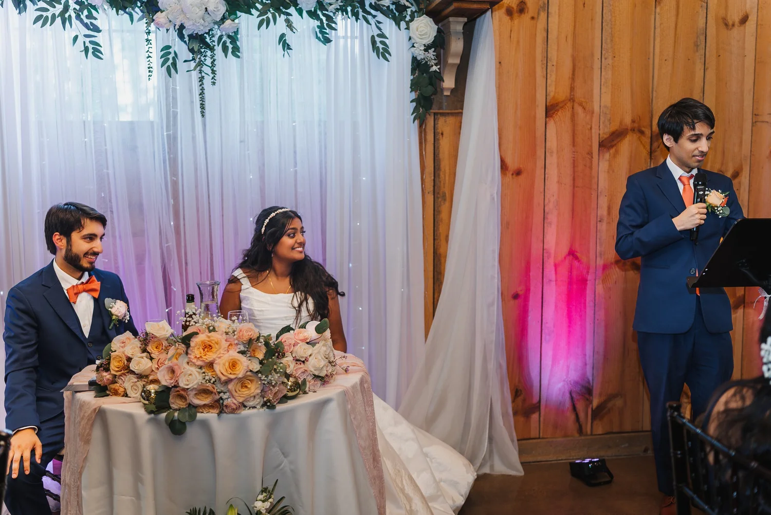 Natalia and Ansel smiling at their sweetheart table while a man gives a toast at the microphone