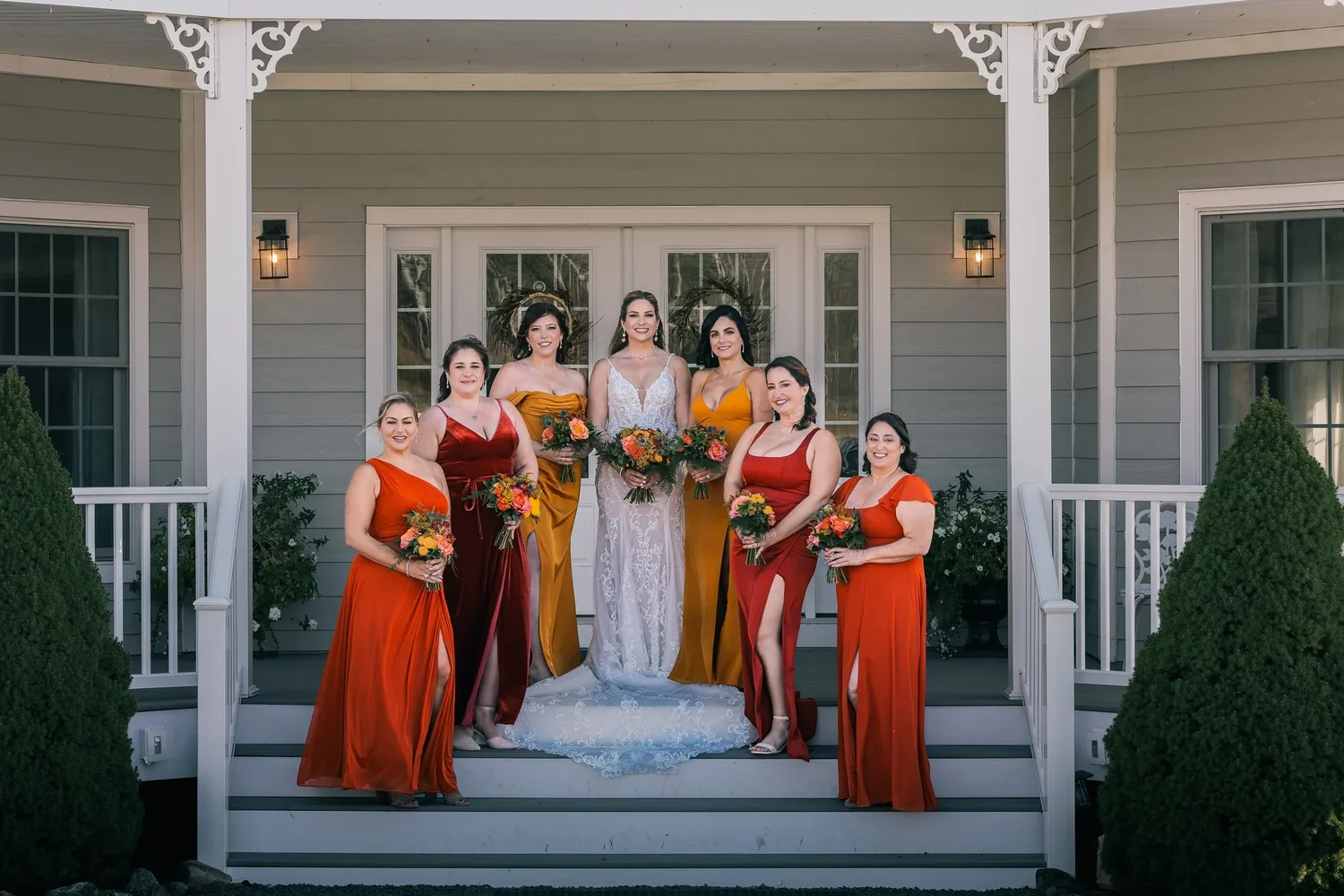 Charissa and her bridesmaids in rust, burnt orange and burgundy gowns on the white porch steps of the Manor House, holding autumn bouquets