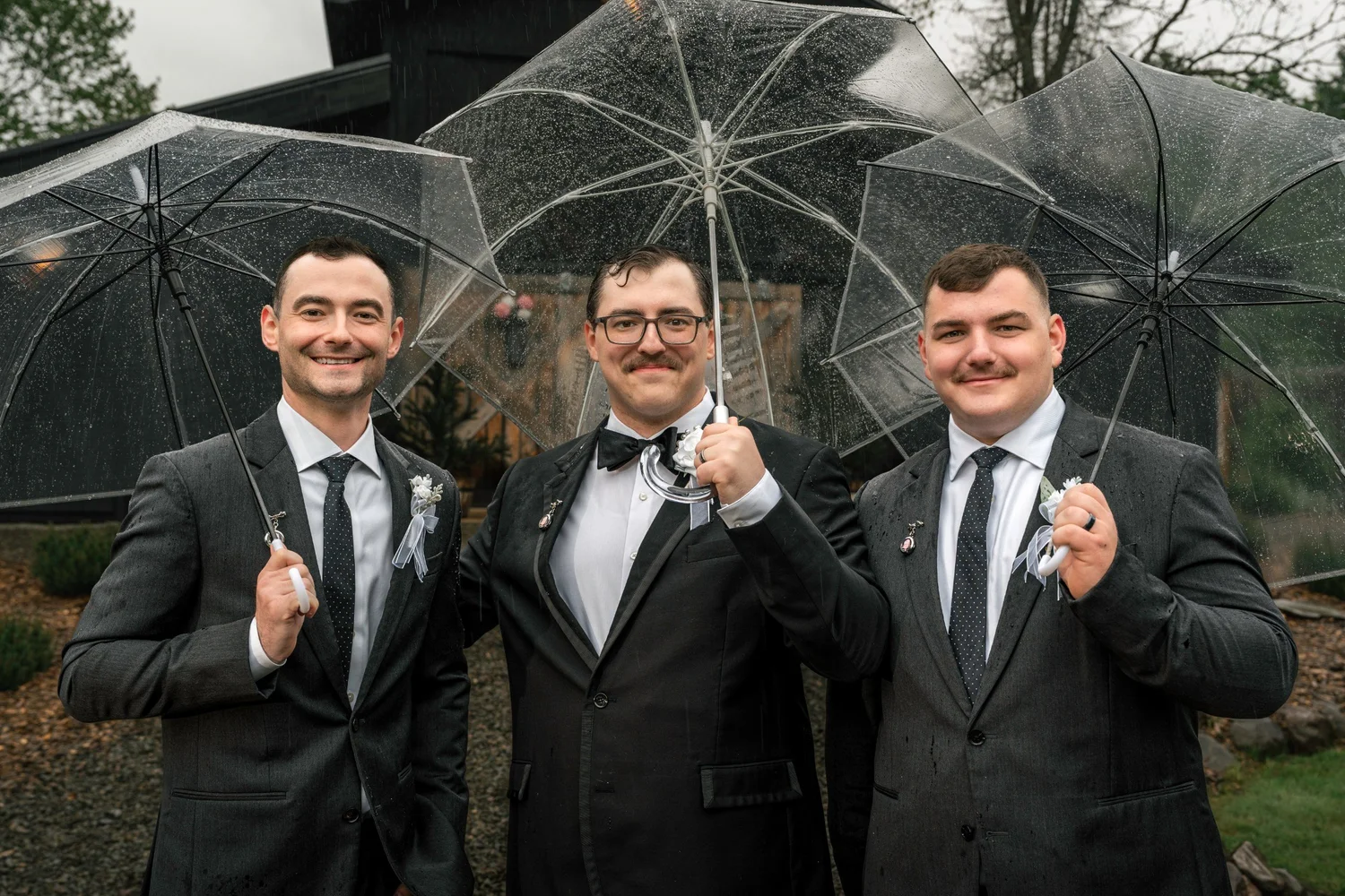 Noah laughing with two groomsmen under clear umbrellas in the rain