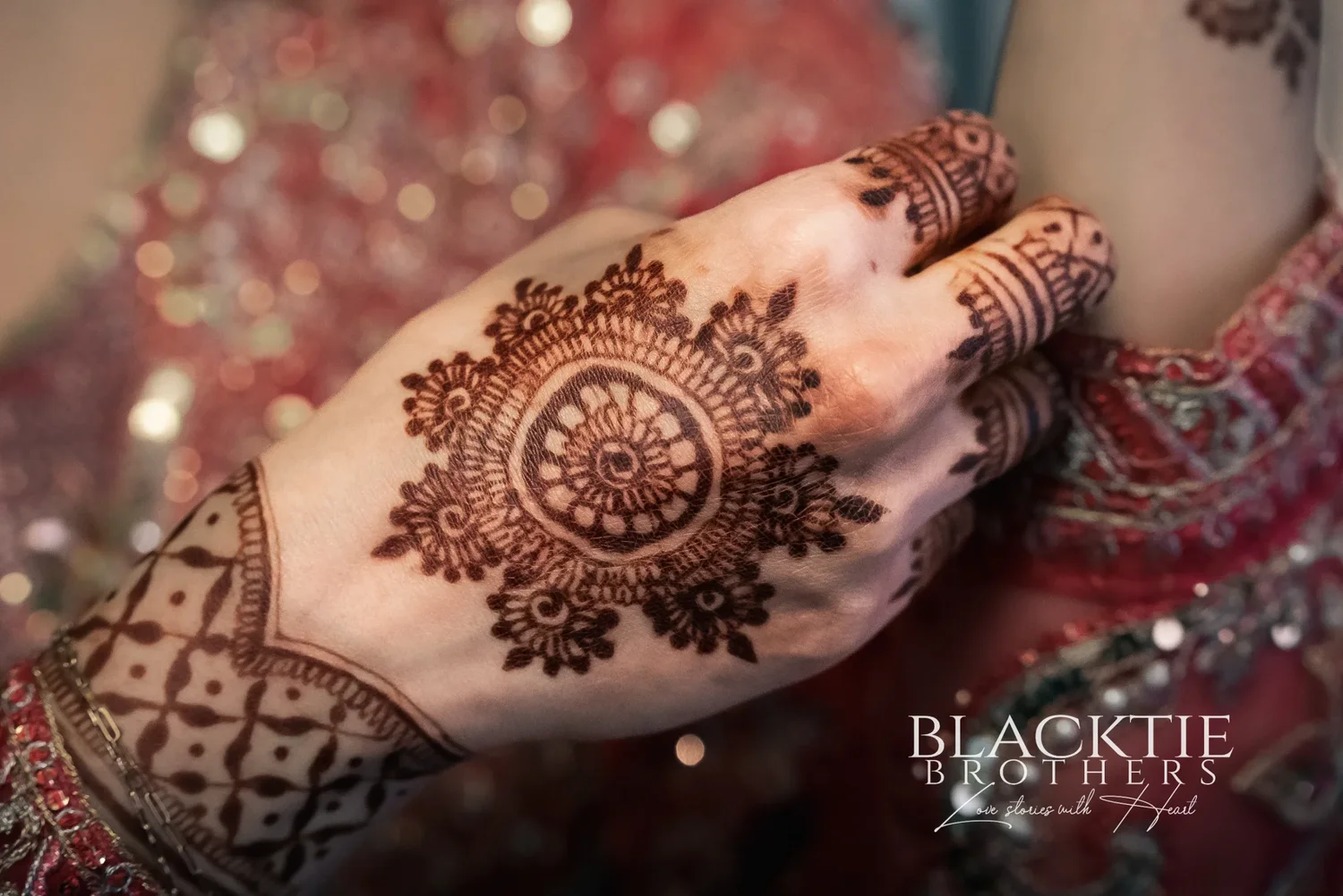 Close view of a hennaed hand with a circular design and patterned fingers, resting against red embroidered fabric