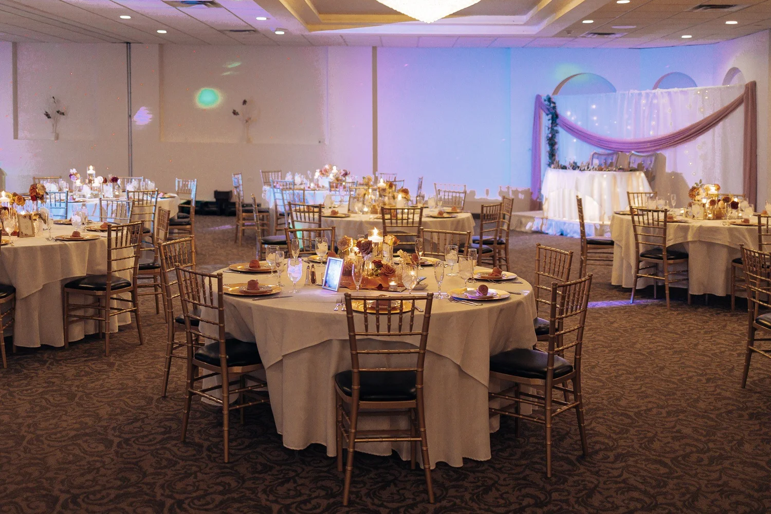 The Riverdale ballroom set for Christina and Erick’s reception, round tables and gold chairs under blush and lavender uplighting