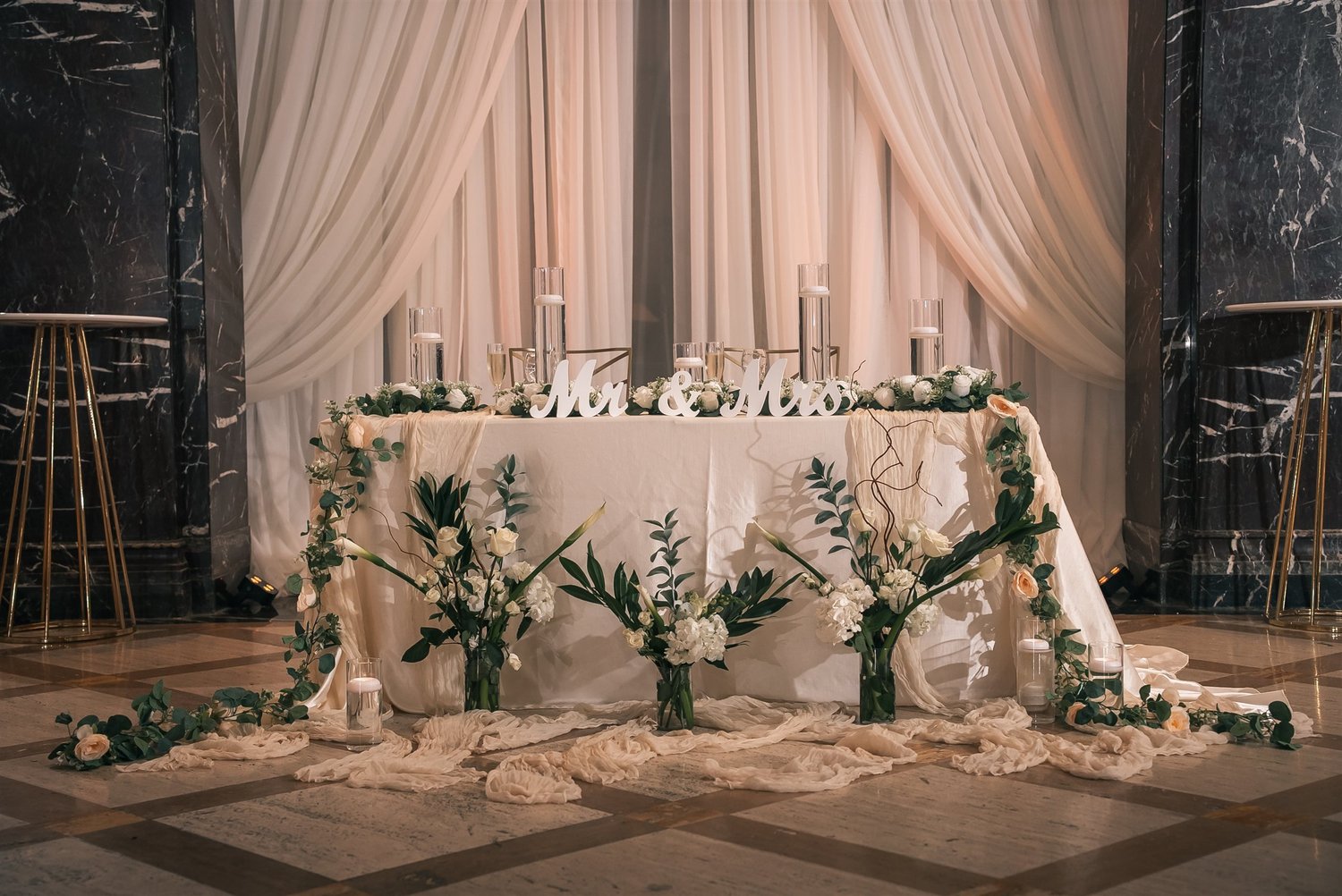 White flowers and greenery surround Tiffany and David’s sweetheart table beneath white draping.