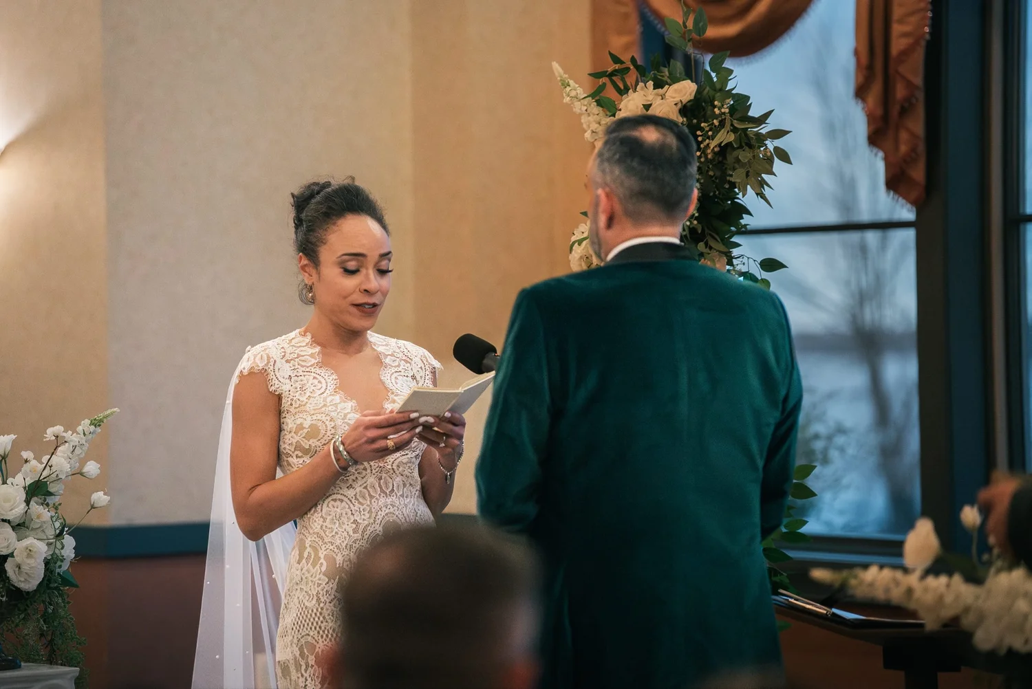 Shannon reading her vows to Leon from a little card, with Seneca Lake just visible through the window behind her