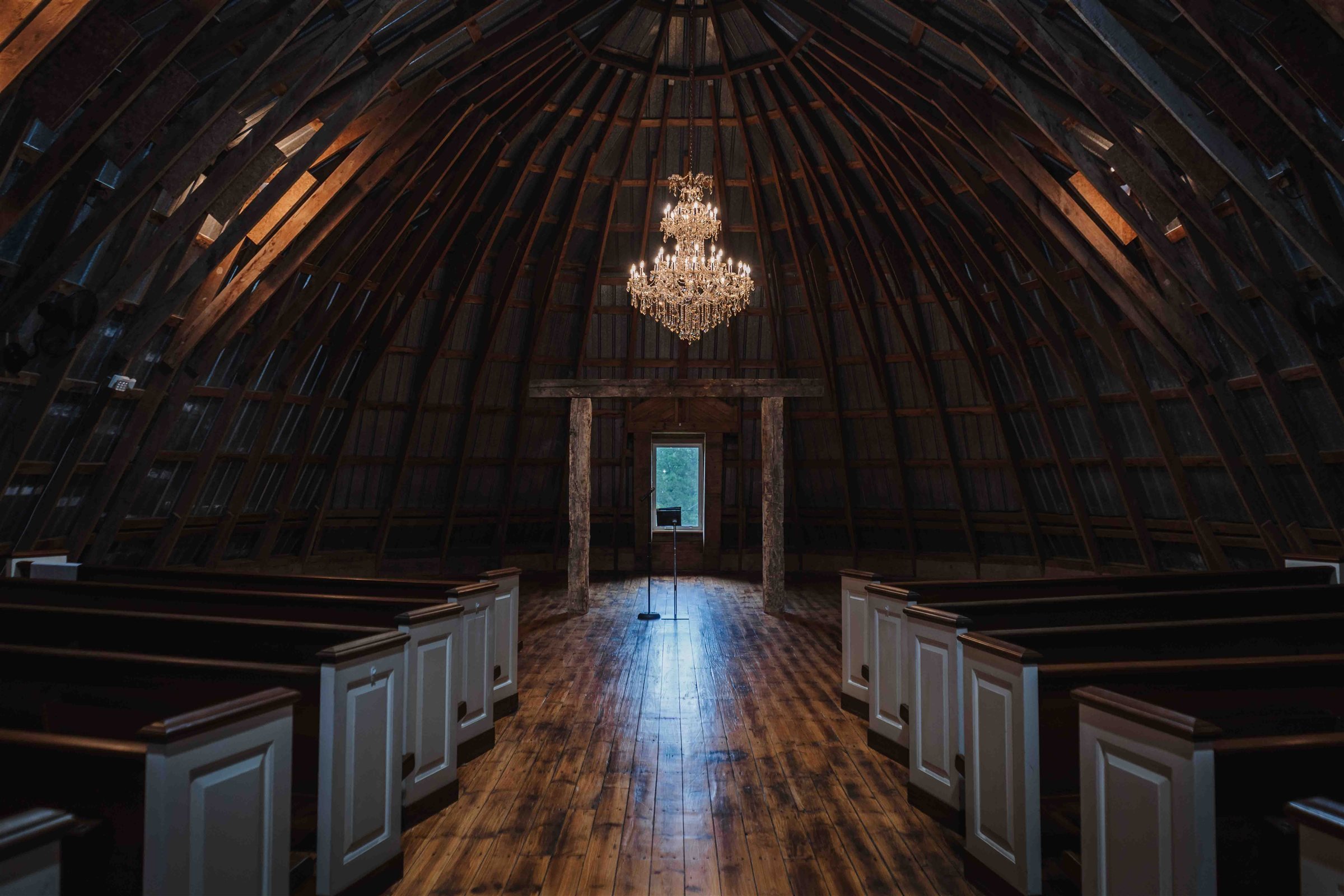 White pews line a wooden aisle beneath exposed barn rafters and a crystal chandelier at The Stone Barn at Beardslee Farm.