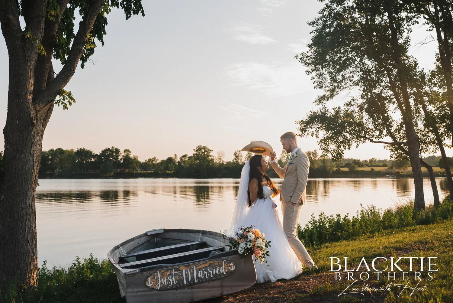 Carolina and Josh at the water's edge at Sally Port View in golden evening light, Josh raising his cowboy hat as she looks up at him, a gray rowboat with a Just Married sign and her bouquet beside them, trees framing the calm water