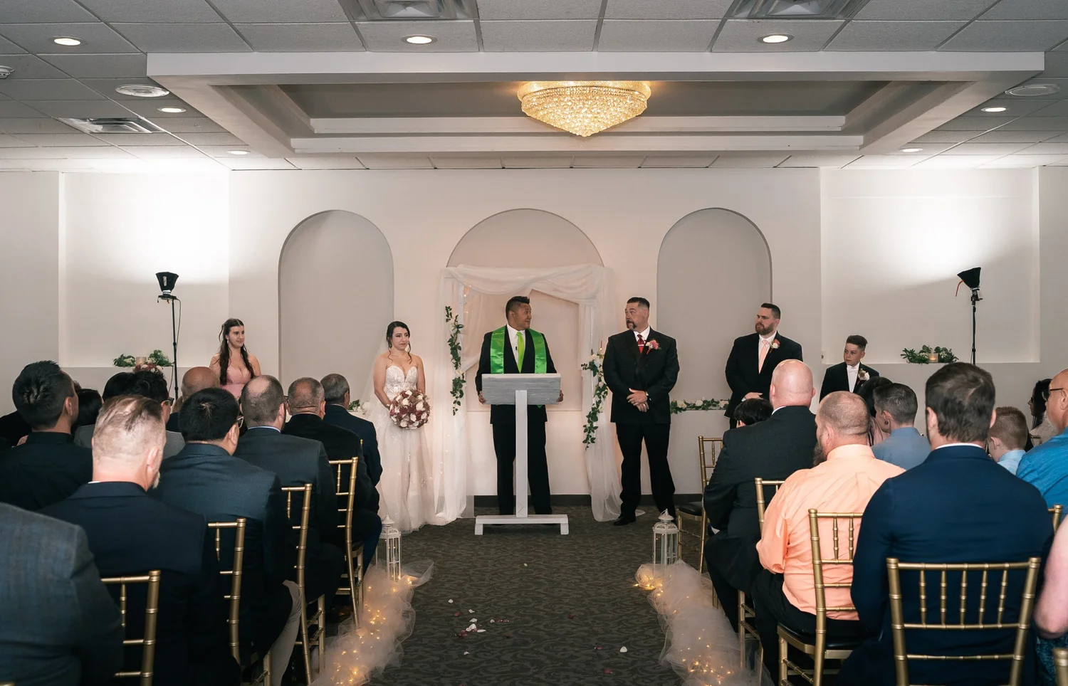 Laura and Dante at the front of the ceremony room with their officiant, framed by white arched alcoves, guests on gold chairs and a chandelier overhead