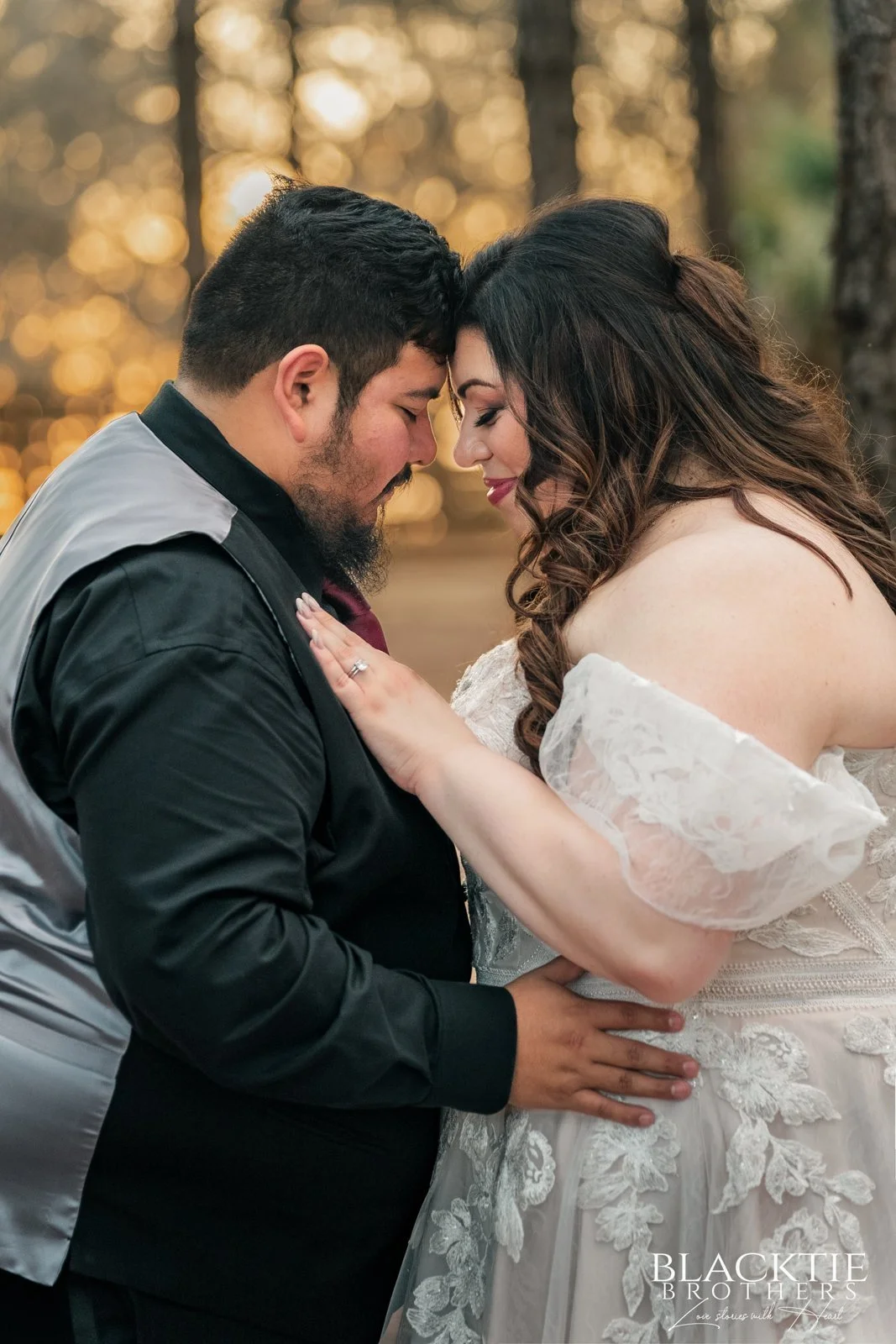 Alyssa and Alberto touch foreheads in a close portrait with warm late-day light filtering through pine trees behind them.