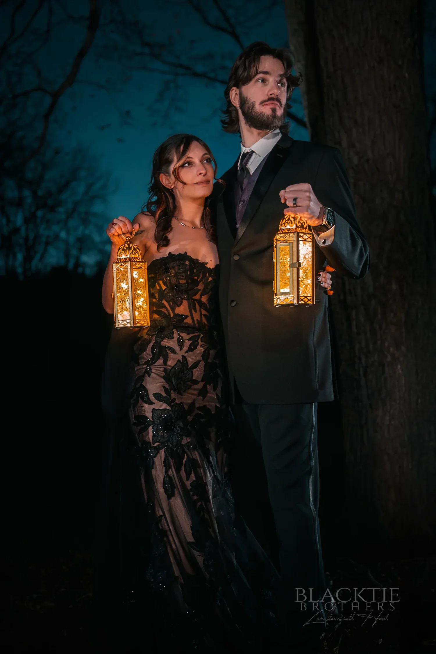 Maddy in a black wedding gown and Dan holding glowing lanterns in the woods at night at their Halloween wedding