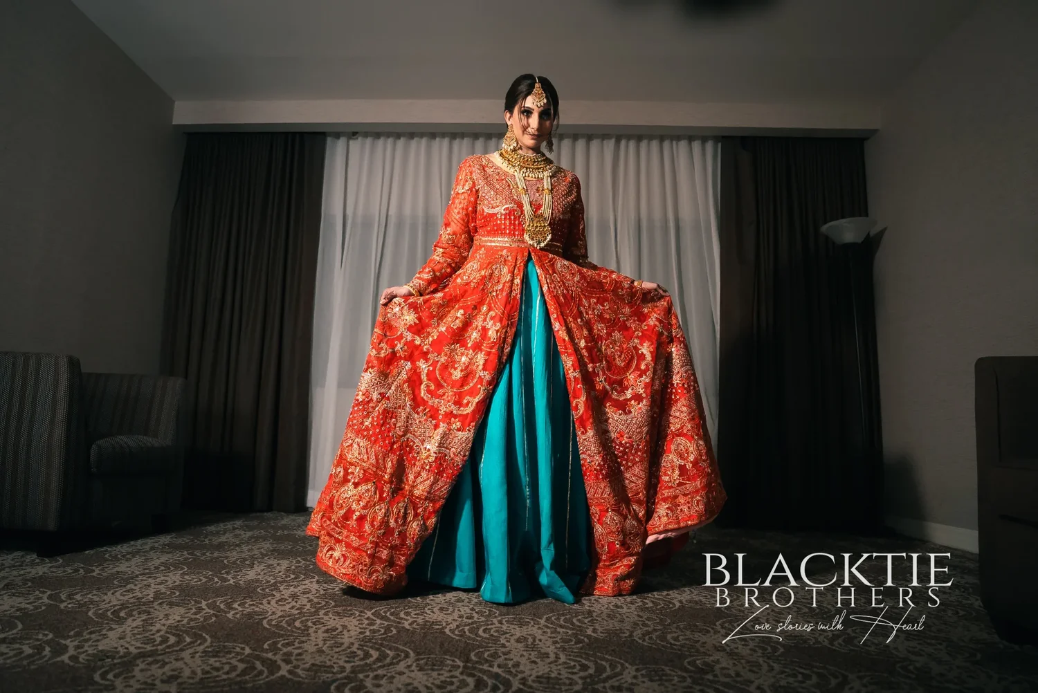 Jia standing in front of the curtained window of her hotel room, holding out the skirt of a red and gold embroidered outfit with a teal panel