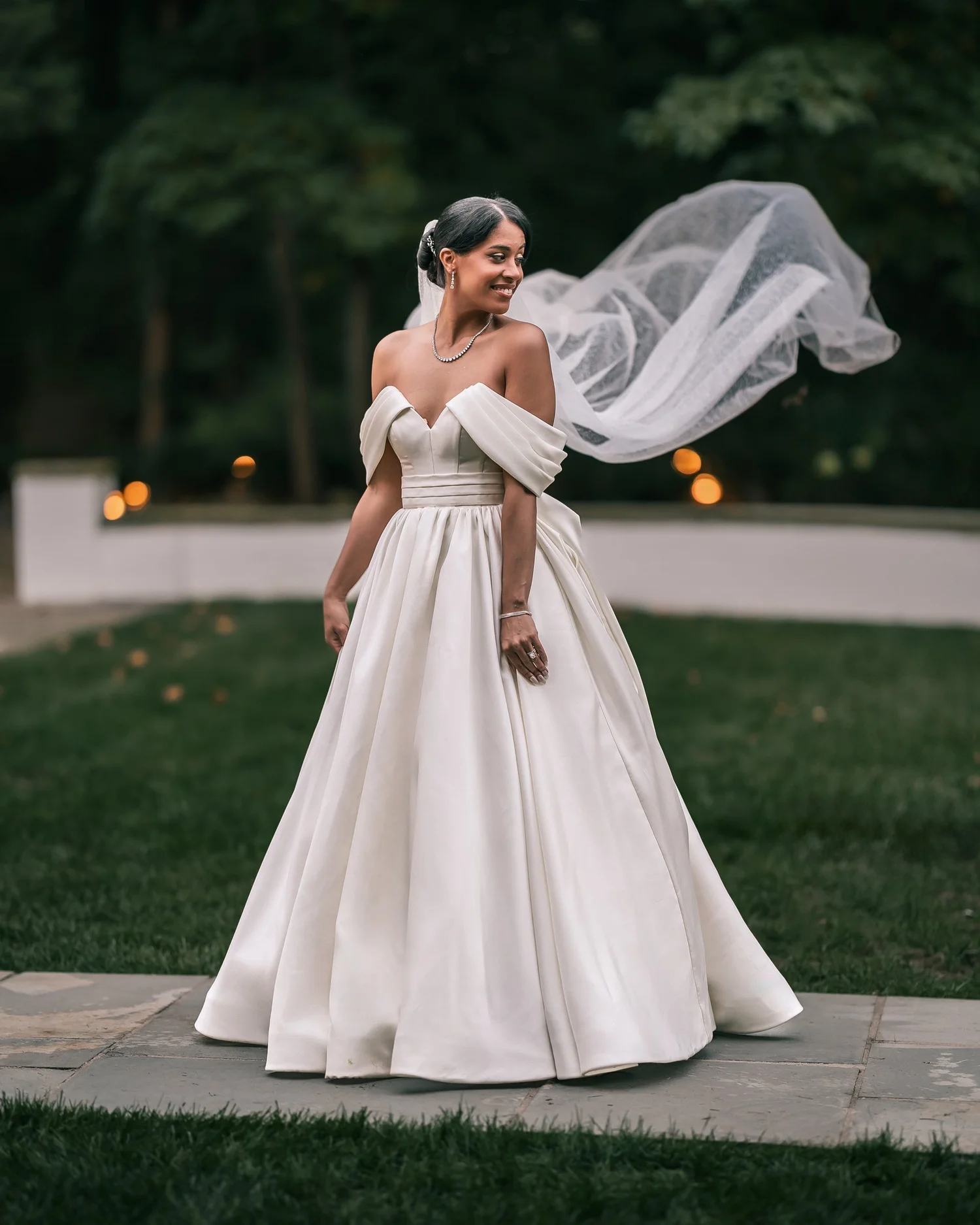 Patricia in an off-the-shoulder ball gown smiles over her shoulder as her veil lifts in the wind on a lawn.