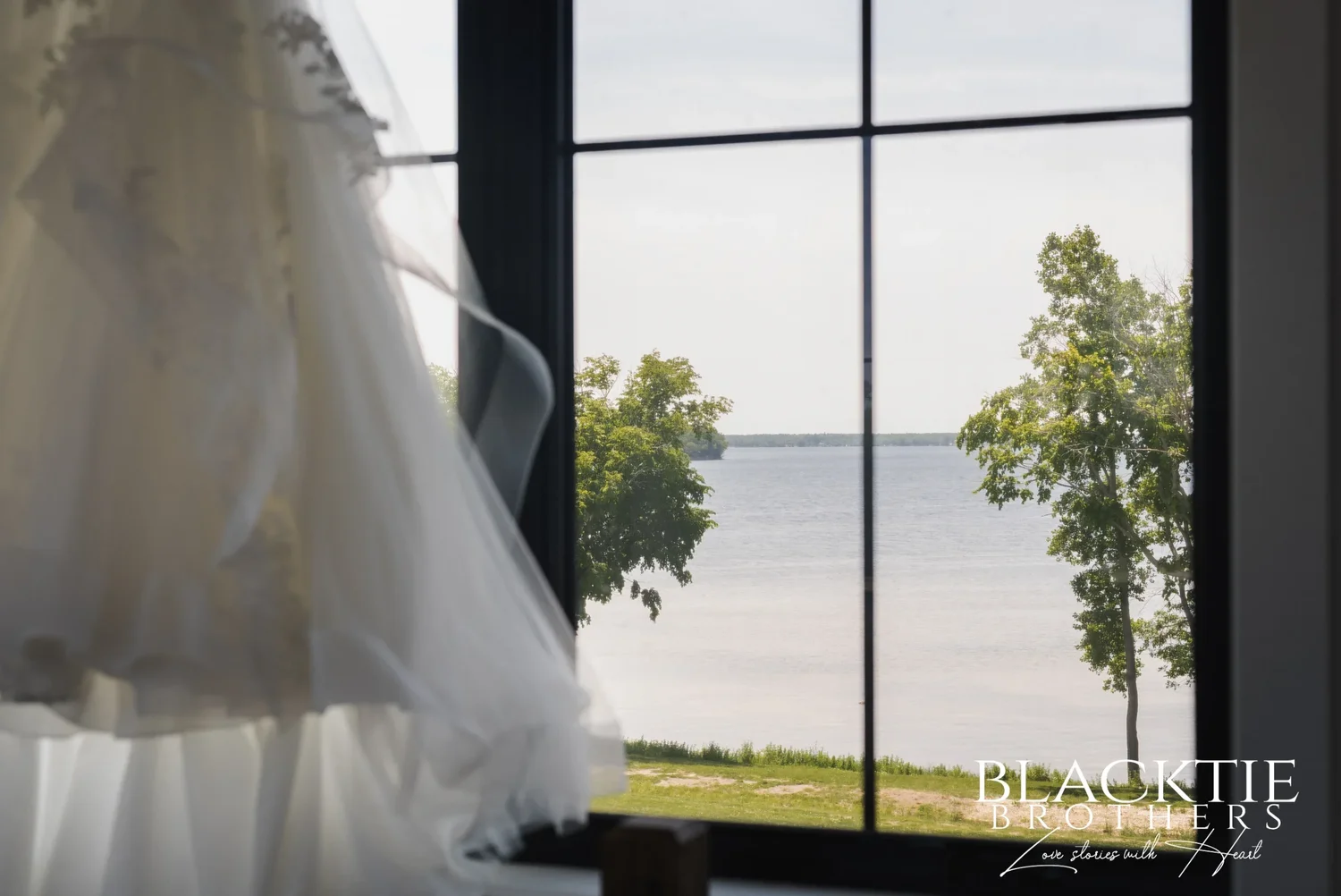 Carolina's wedding dress hanging in a tall black-framed window upstairs at Sally Port View, the water and two trees visible through the glass