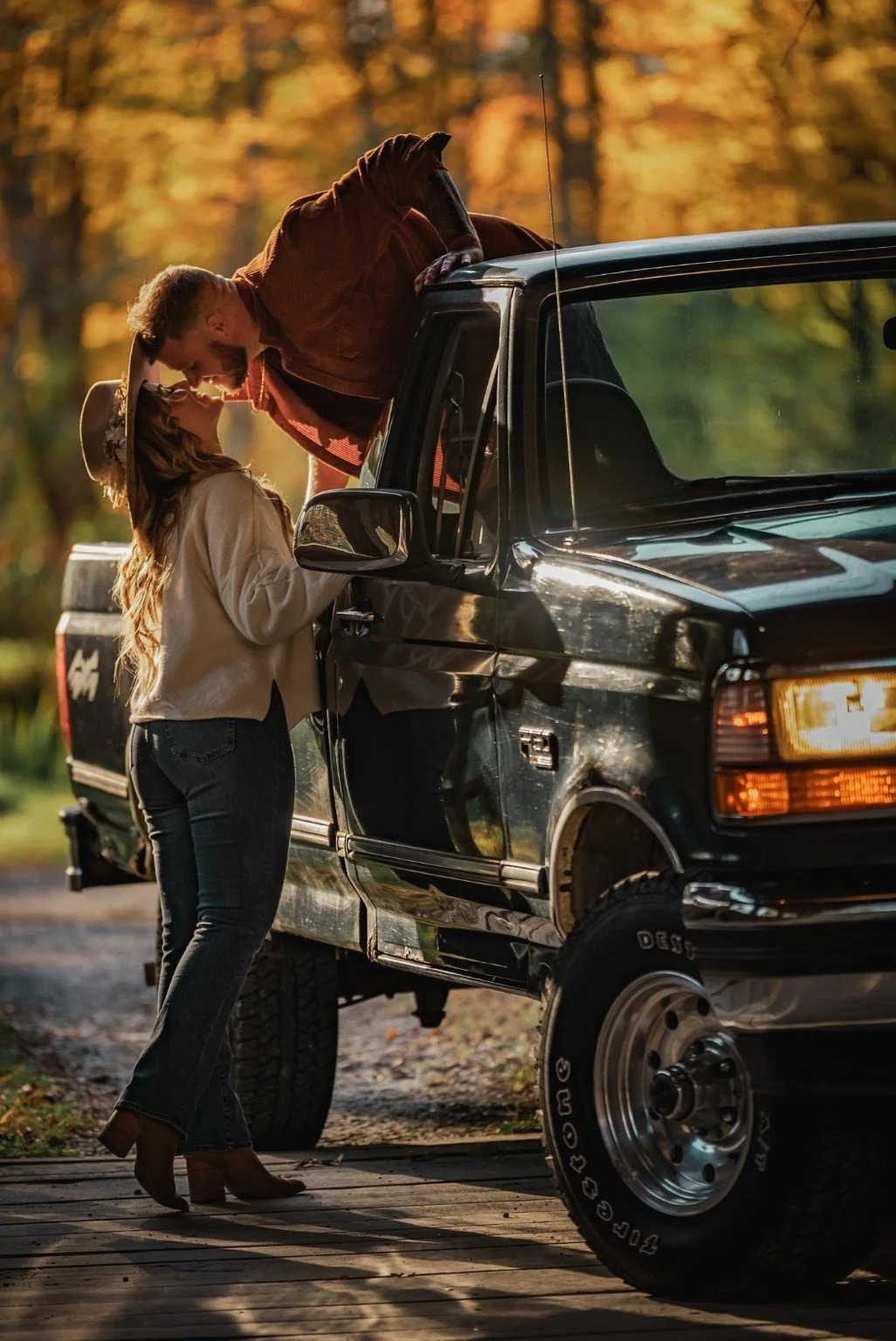A couple kisses beside a dark pickup truck, with golden autumn leaves behind them.