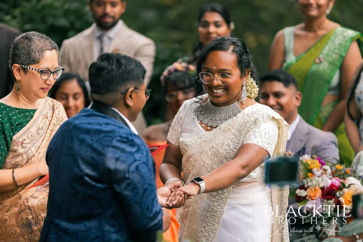 Hima and Alina holding hands and smiling at each other during the ceremony, guests and the wedding party watching around them