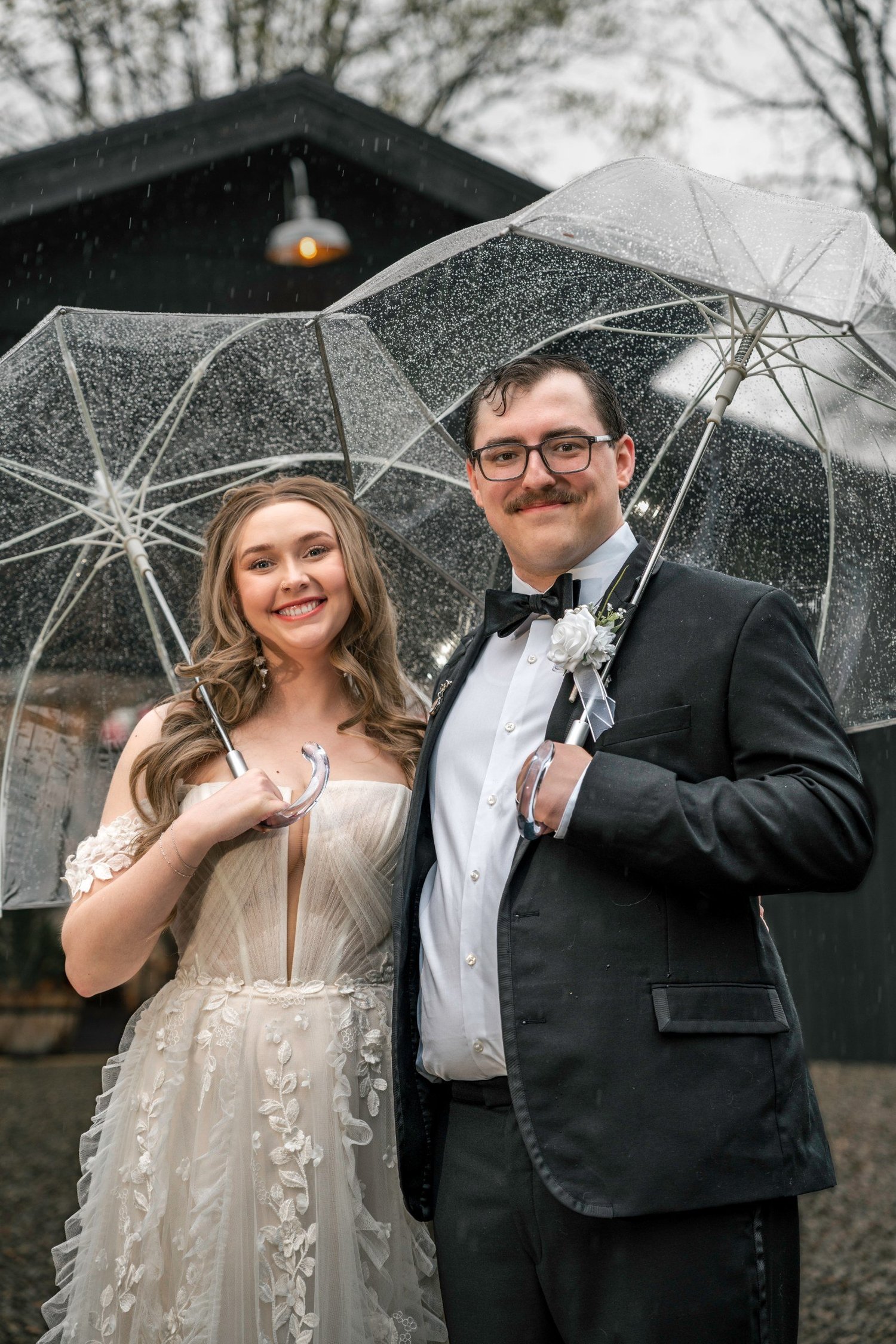 Lindsey and Noah smiling under clear umbrellas in front of the Black Barn