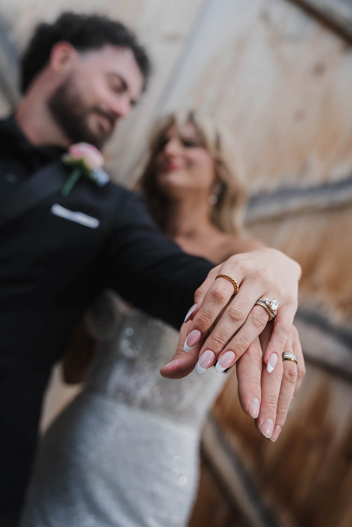Kari’s hand rests on Josh’s, showing her engagement ring and band and his wedding band, with the couple softly out of focus behind.