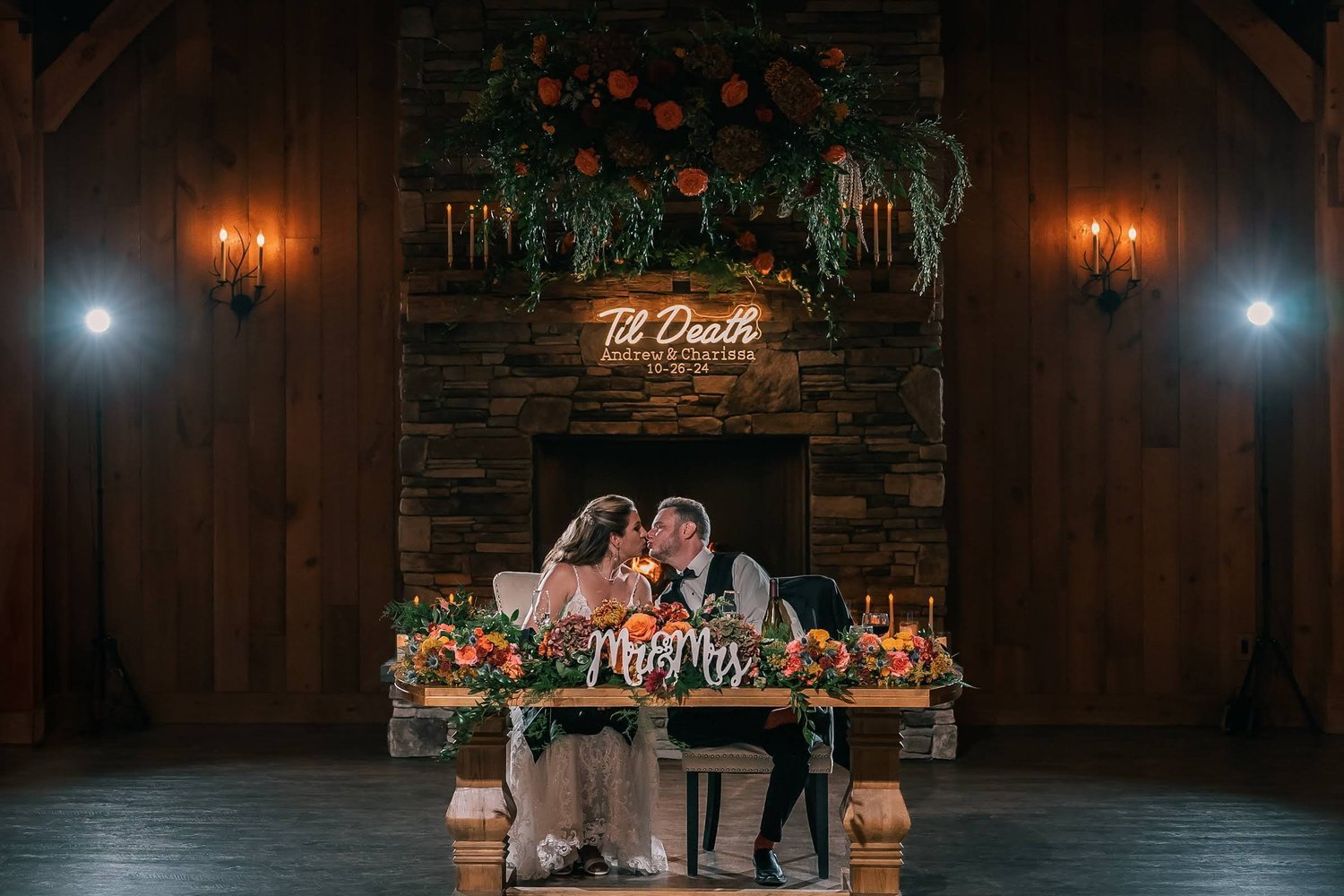 Charissa and Andrew kissing at their sweetheart table in front of the Barn's grand stone fireplace, a neon Til Death sign and autumn garland above them