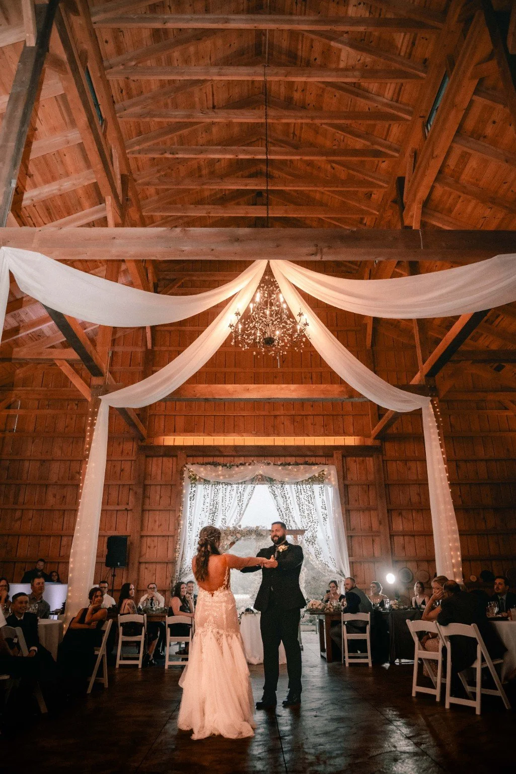 Joelle and Louie share their first dance beneath a chandelier, white draping and the barn’s exposed timber beams.