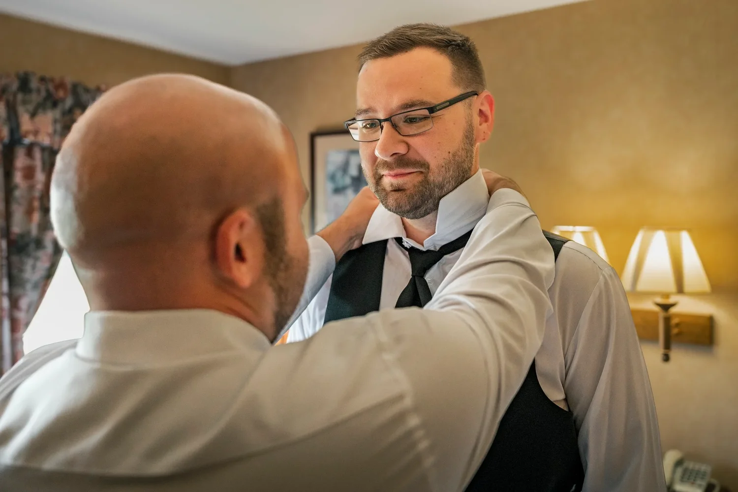 Zach in a vest and glasses while a groomsman straightens his tie in a hotel room at Traditions