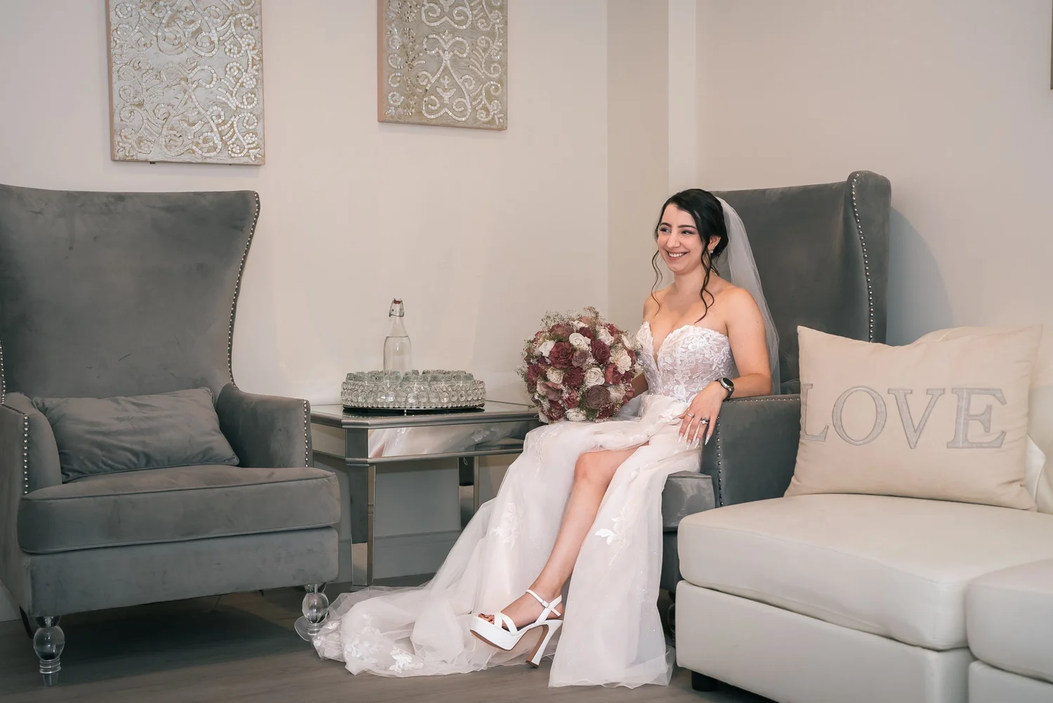 Laura sitting in the bridal suite at The Riverdale with a bouquet of burgundy and blush roses, gray wingback chairs and a white couch with a LOVE pillow beside her