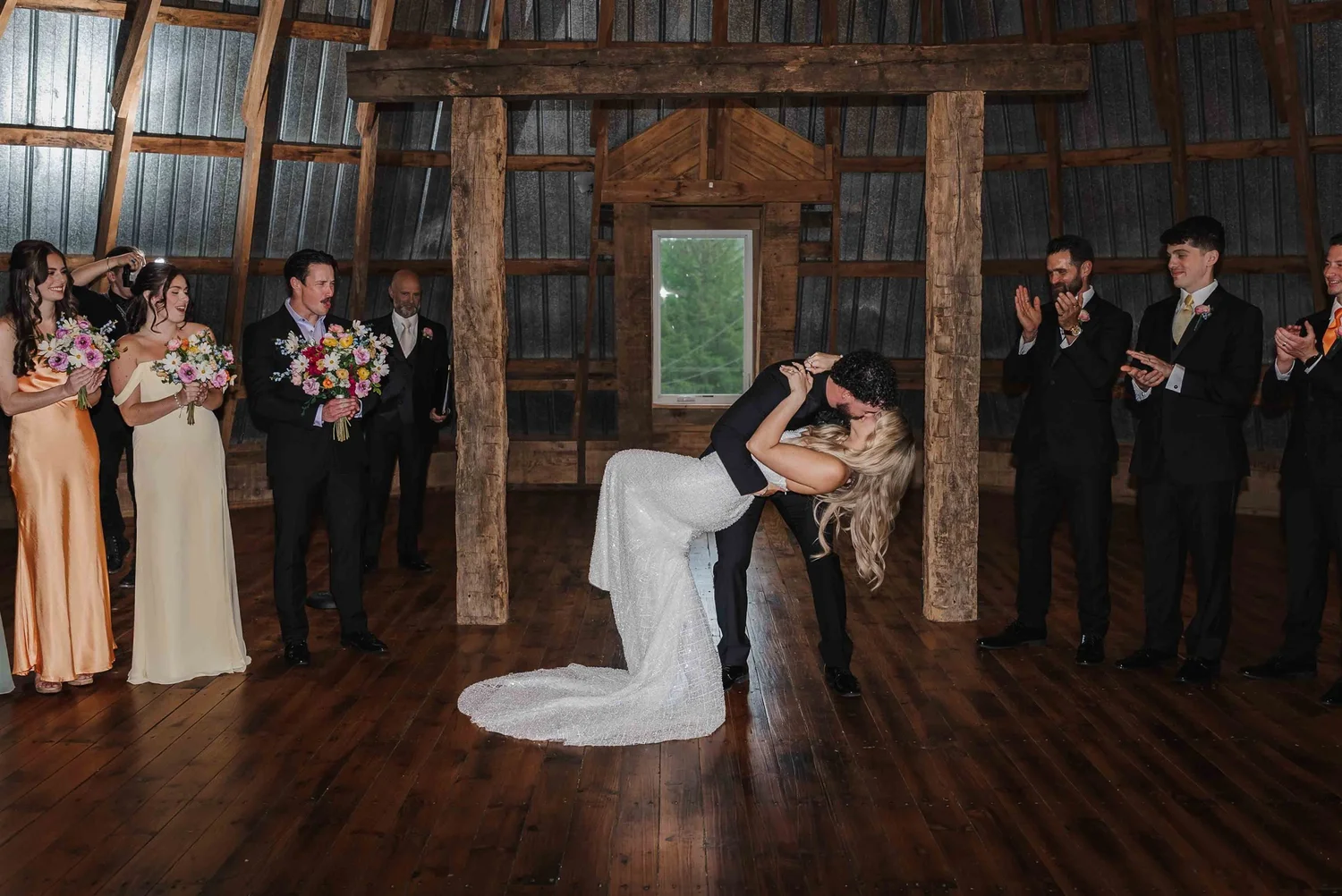 Josh dips Kari for a kiss while their wedding party applauds inside the barn.