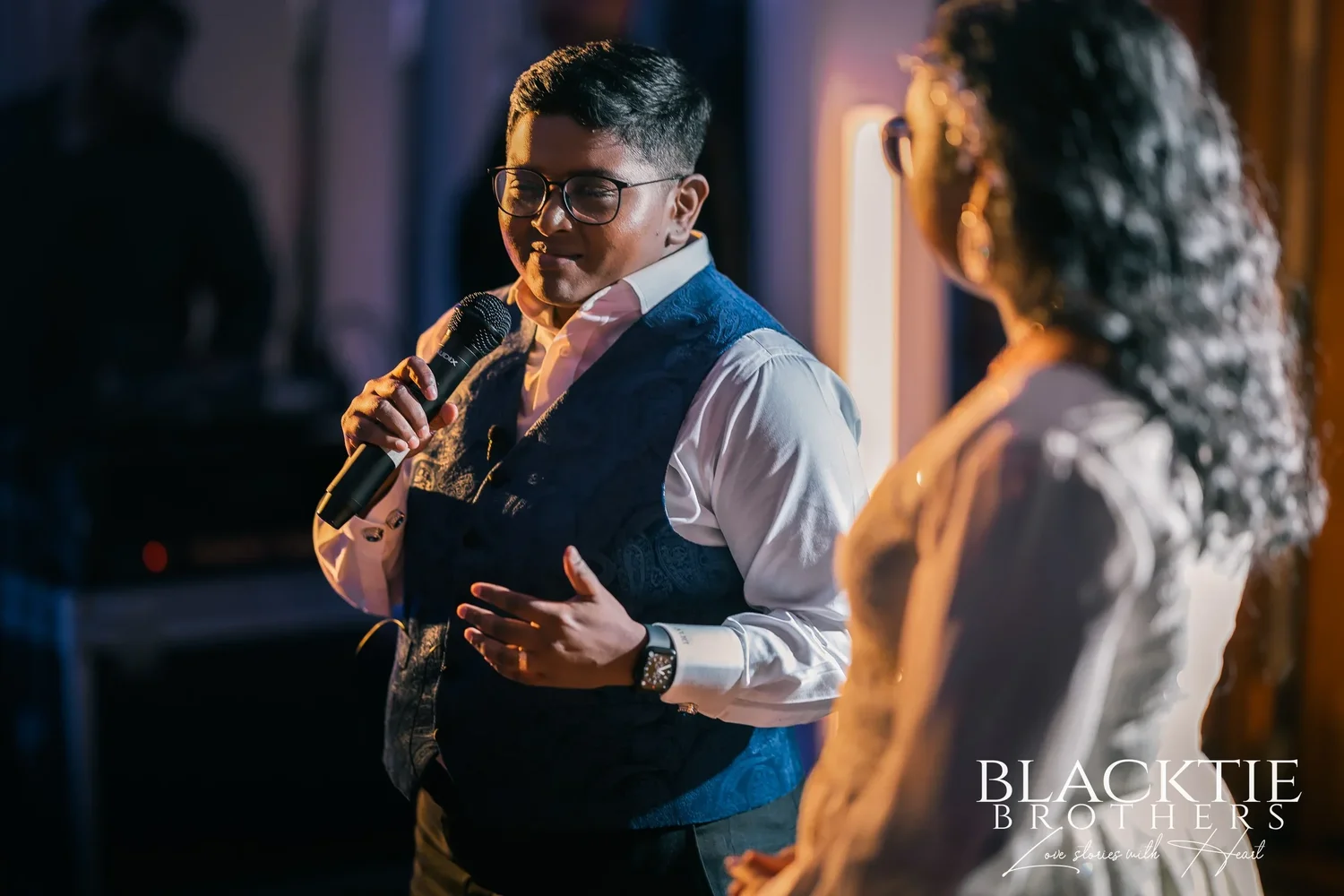 One of the couple giving a speech into a microphone in the evening, lit warmly against the dark tent