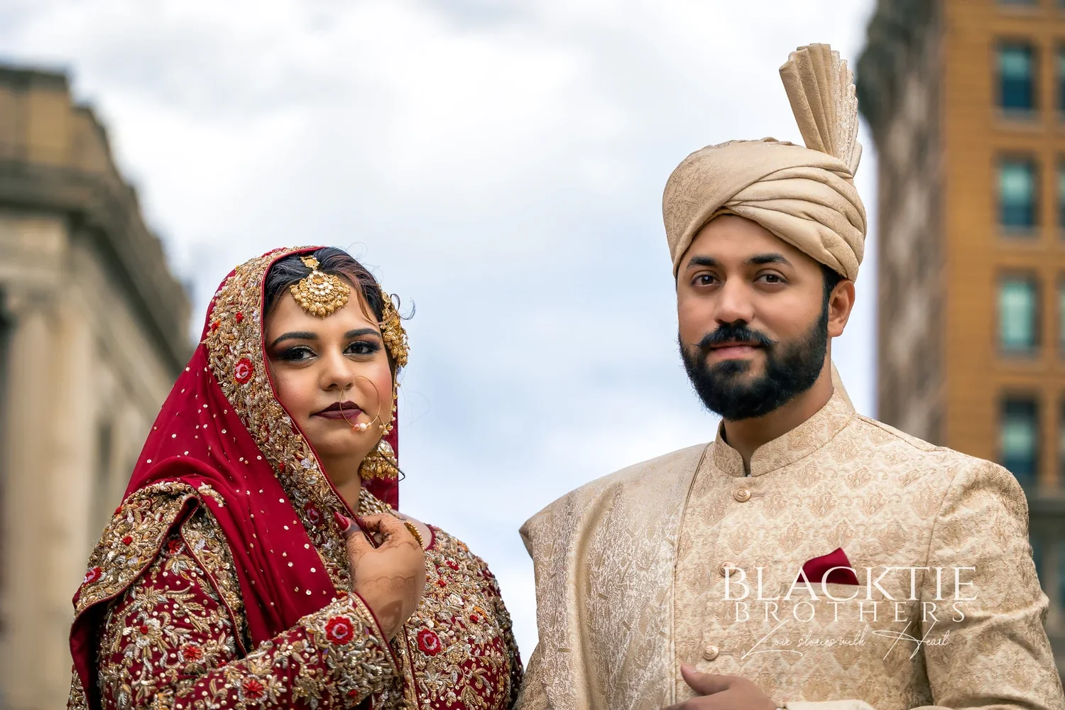 Close portrait of the bride in a red veil and gold jewelry and the groom in an ivory turban, city buildings and an overcast sky behind them