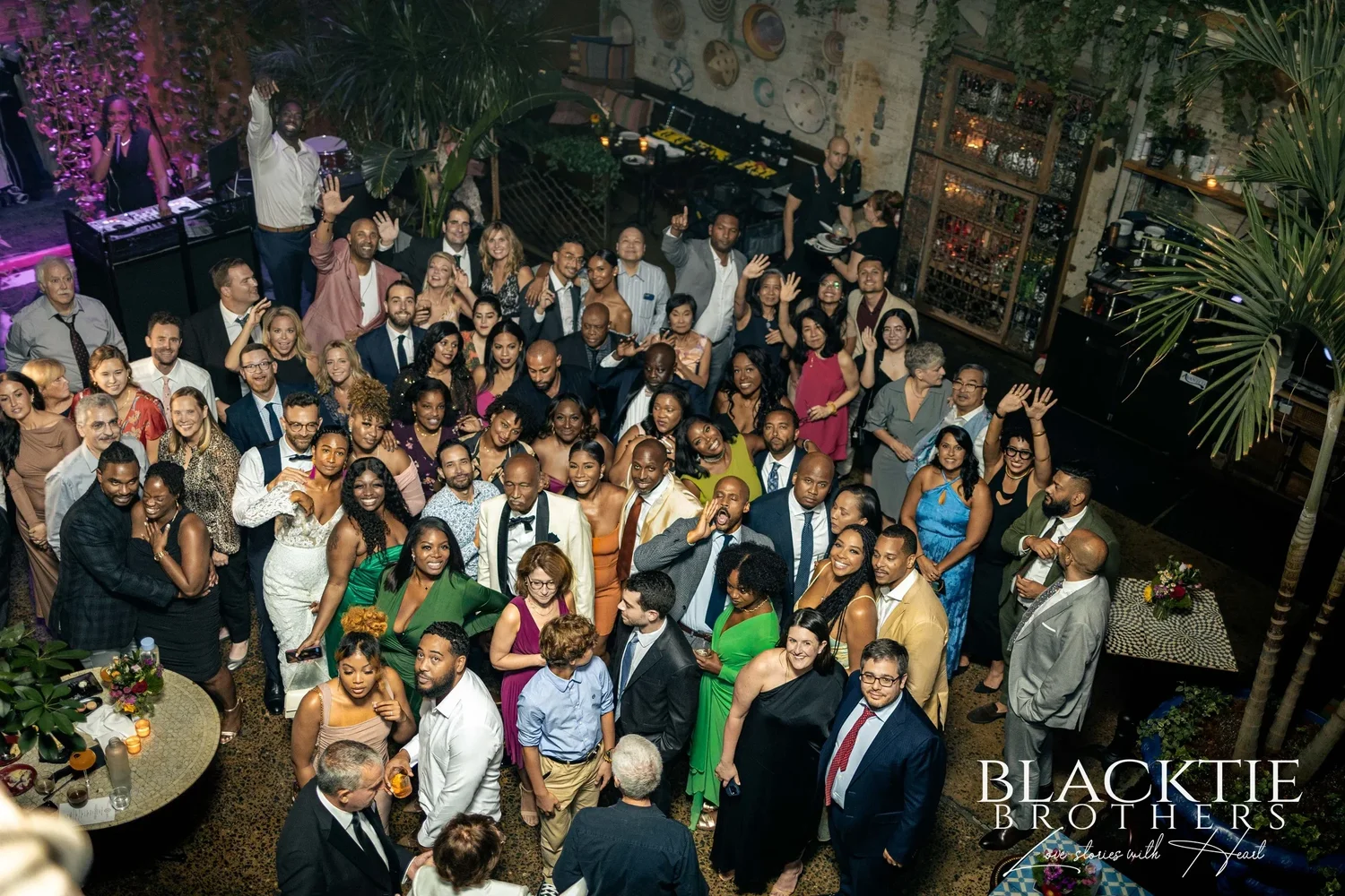 The whole wedding crowd photographed from the mezzanine, dozens of guests looking up and waving from the floor of the main room