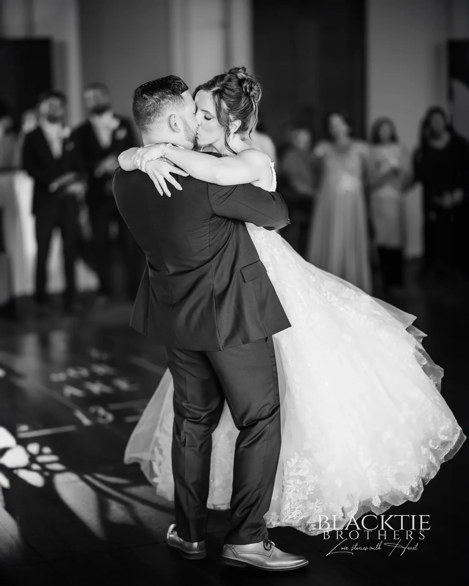 Black and white photograph of Taylor and Jake kissing during their first dance, her gown swirling, guests watching from the edge of the dance floor