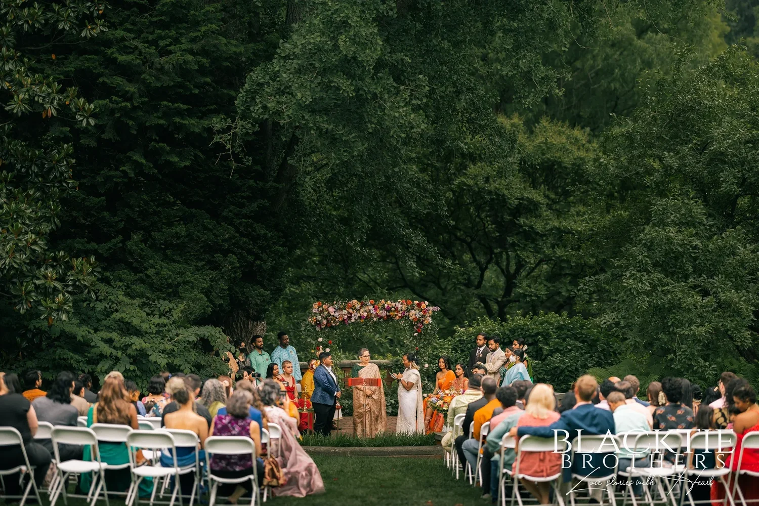 The ceremony at Morris Arboretum seen from the back of the lawn: rows of guests on folding chairs facing the couple and officiant under a floral arch, tall trees all around