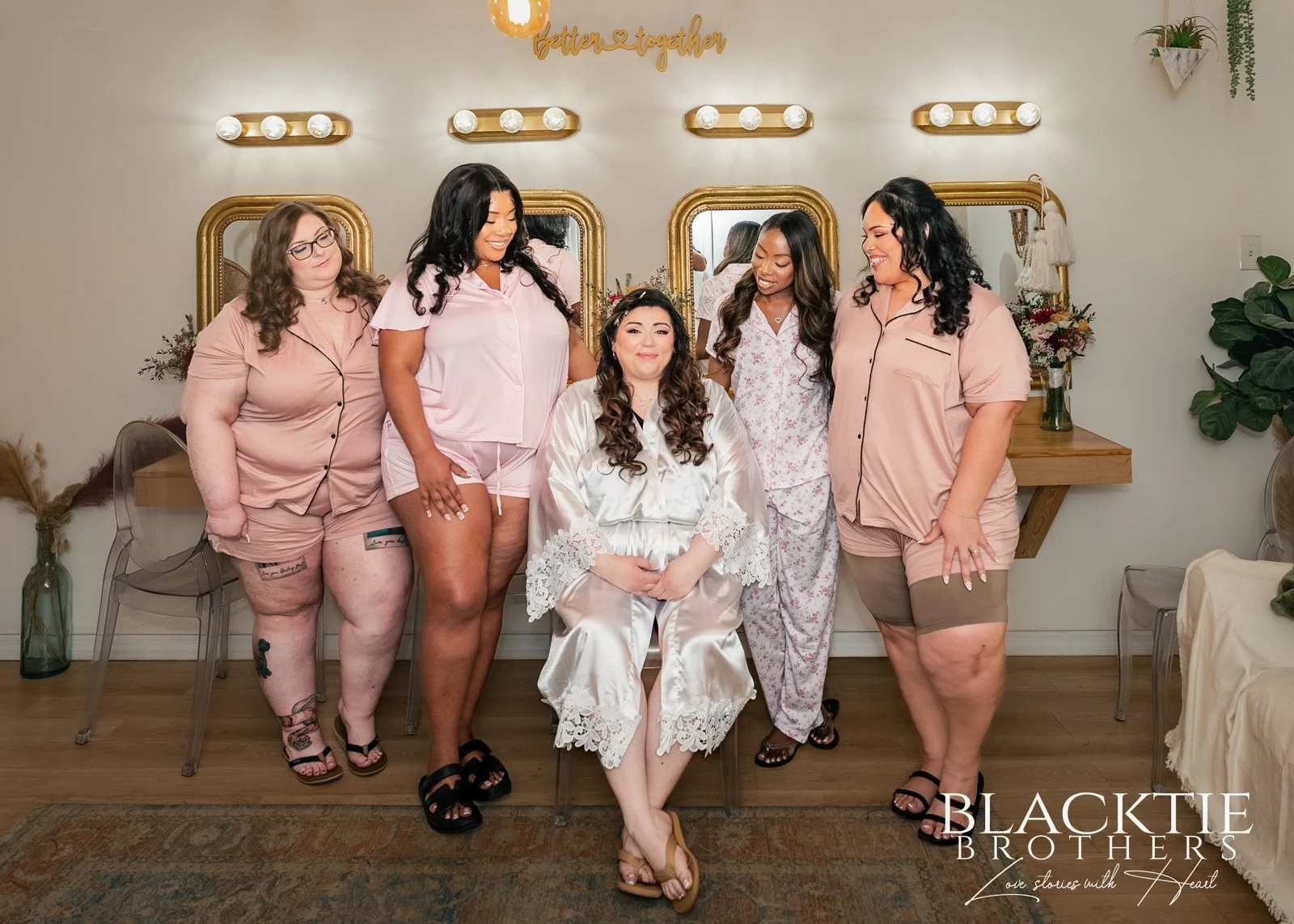 Alyssa sits in a white robe with members of her wedding party in pink pajamas beside illuminated vanity mirrors.