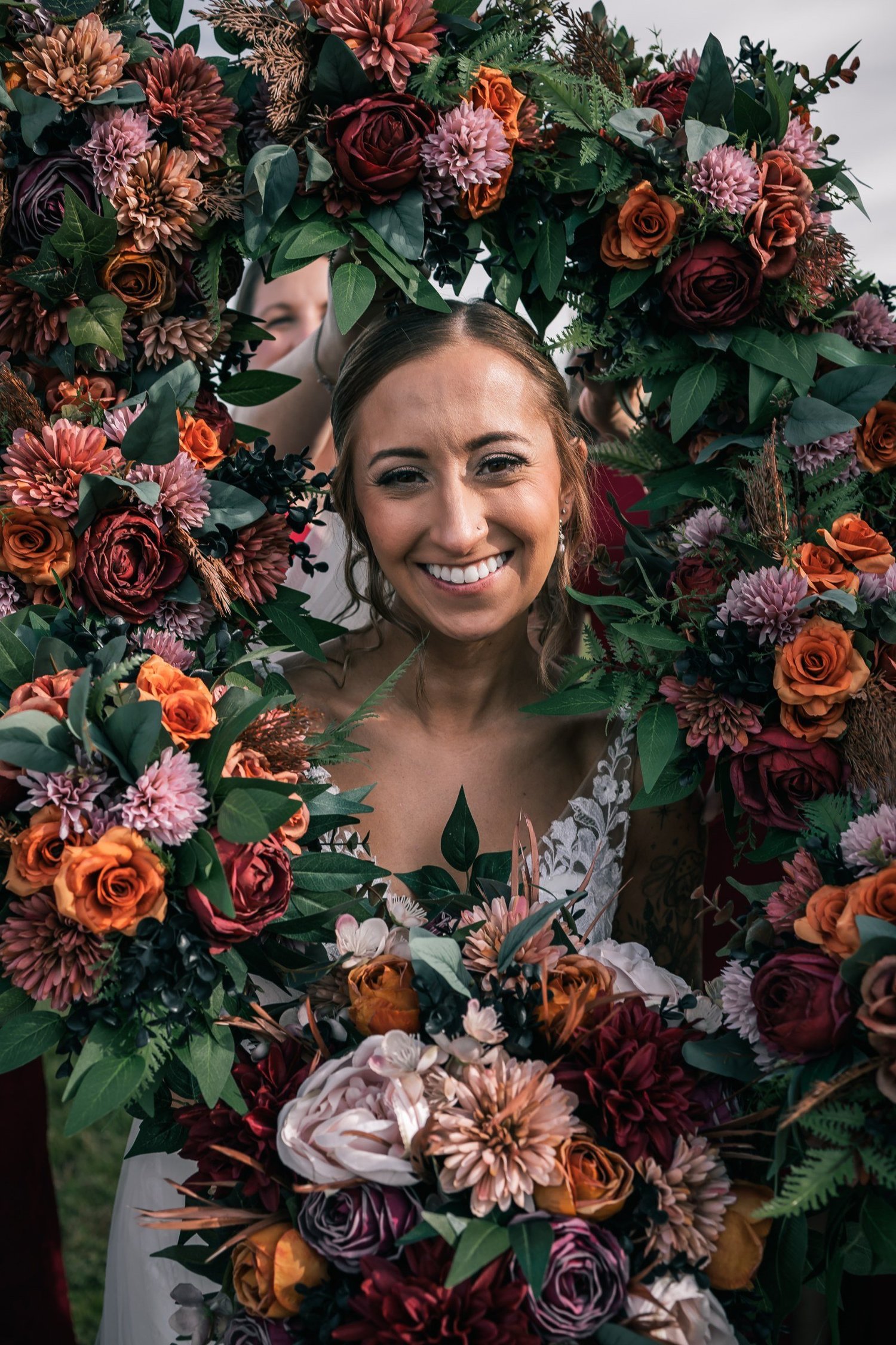Heather smiling through a large round wreath of red, orange and cream flowers