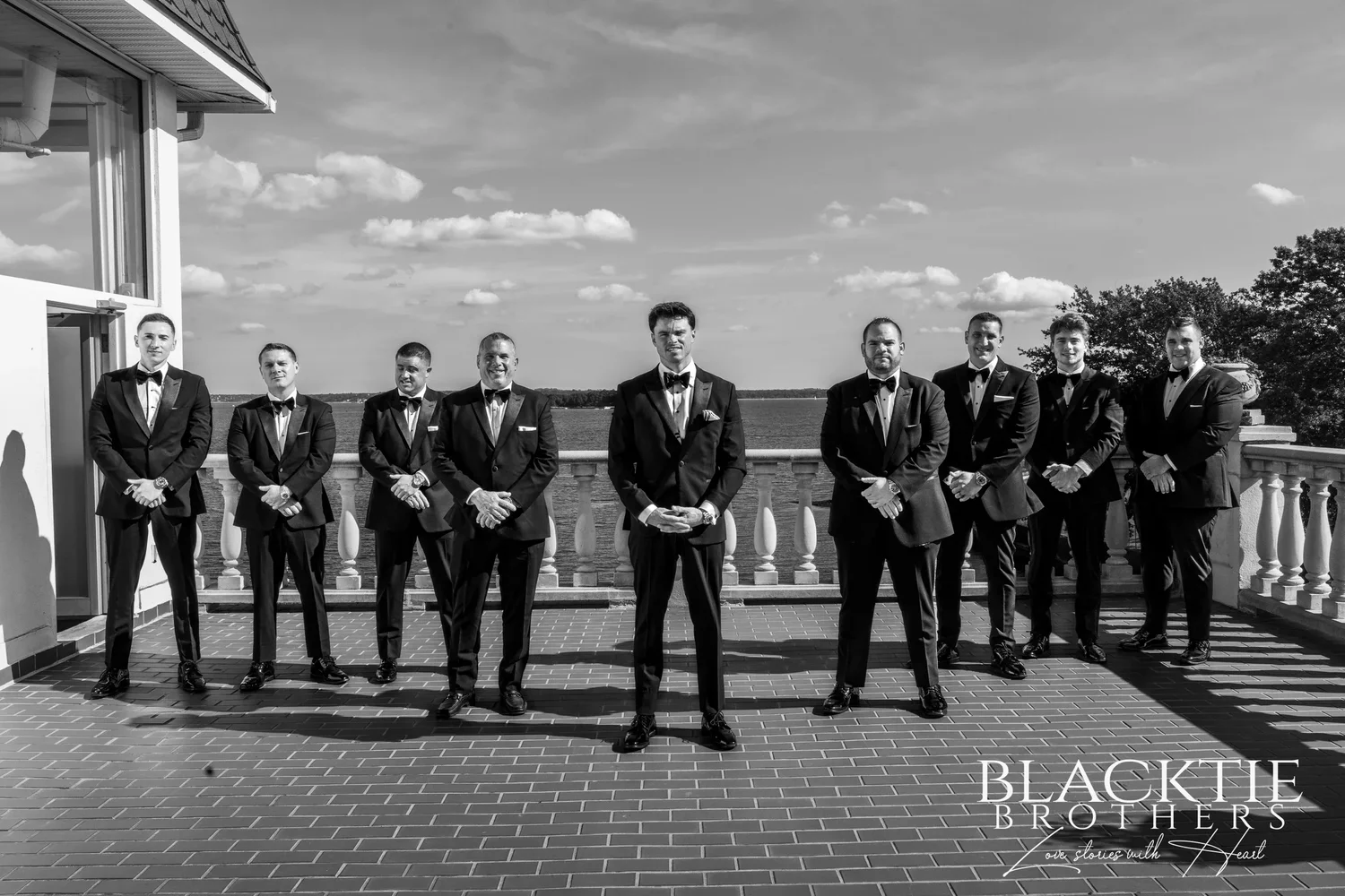 Black and white photograph of the groom's side in tuxedos standing in a line on the brick terrace, the balustrade and Long Island Sound behind them