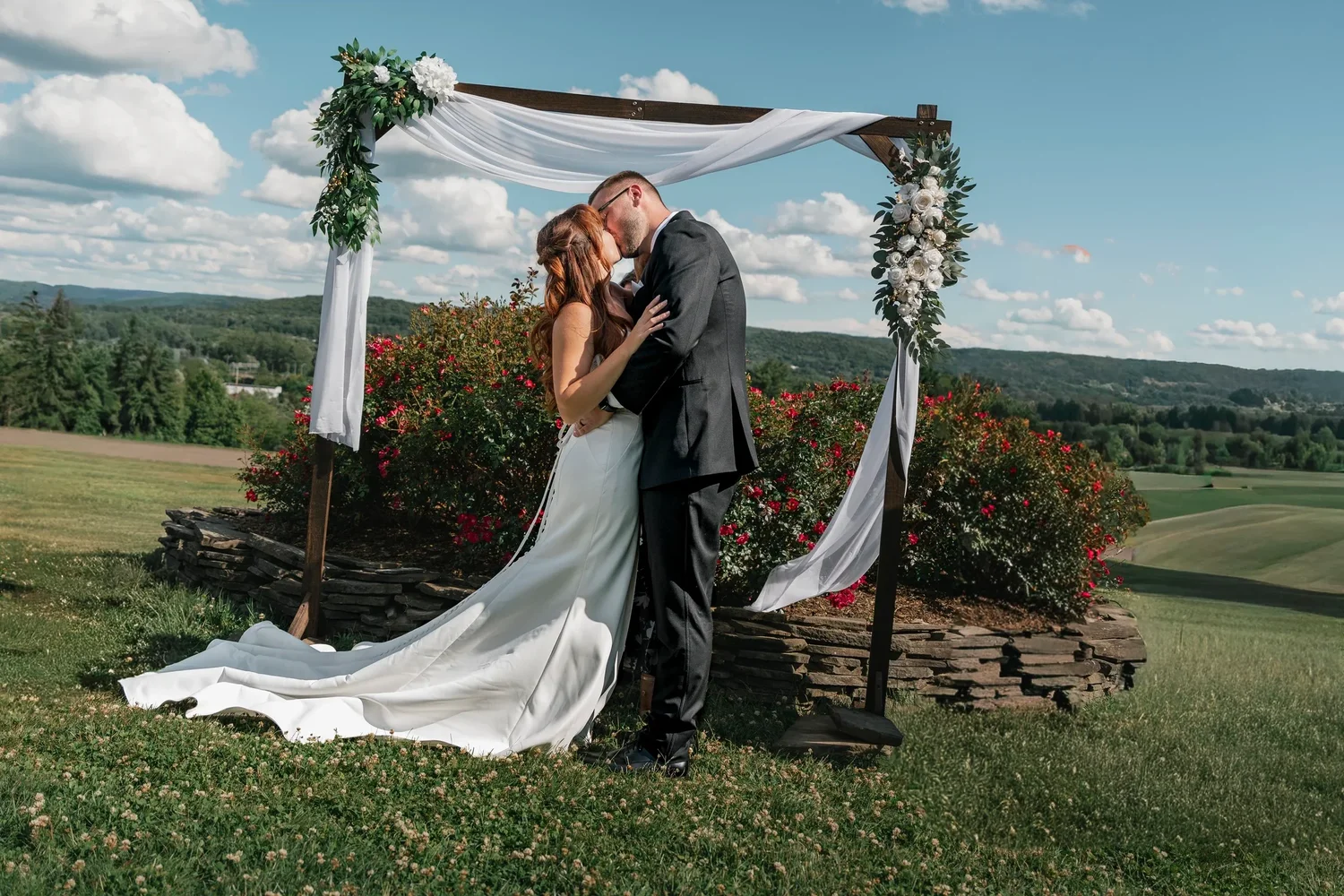 Danielle and Zach kissing beneath a draped wooden arbor at Traditions at the Glen, her train spread across the grass and the hills behind them