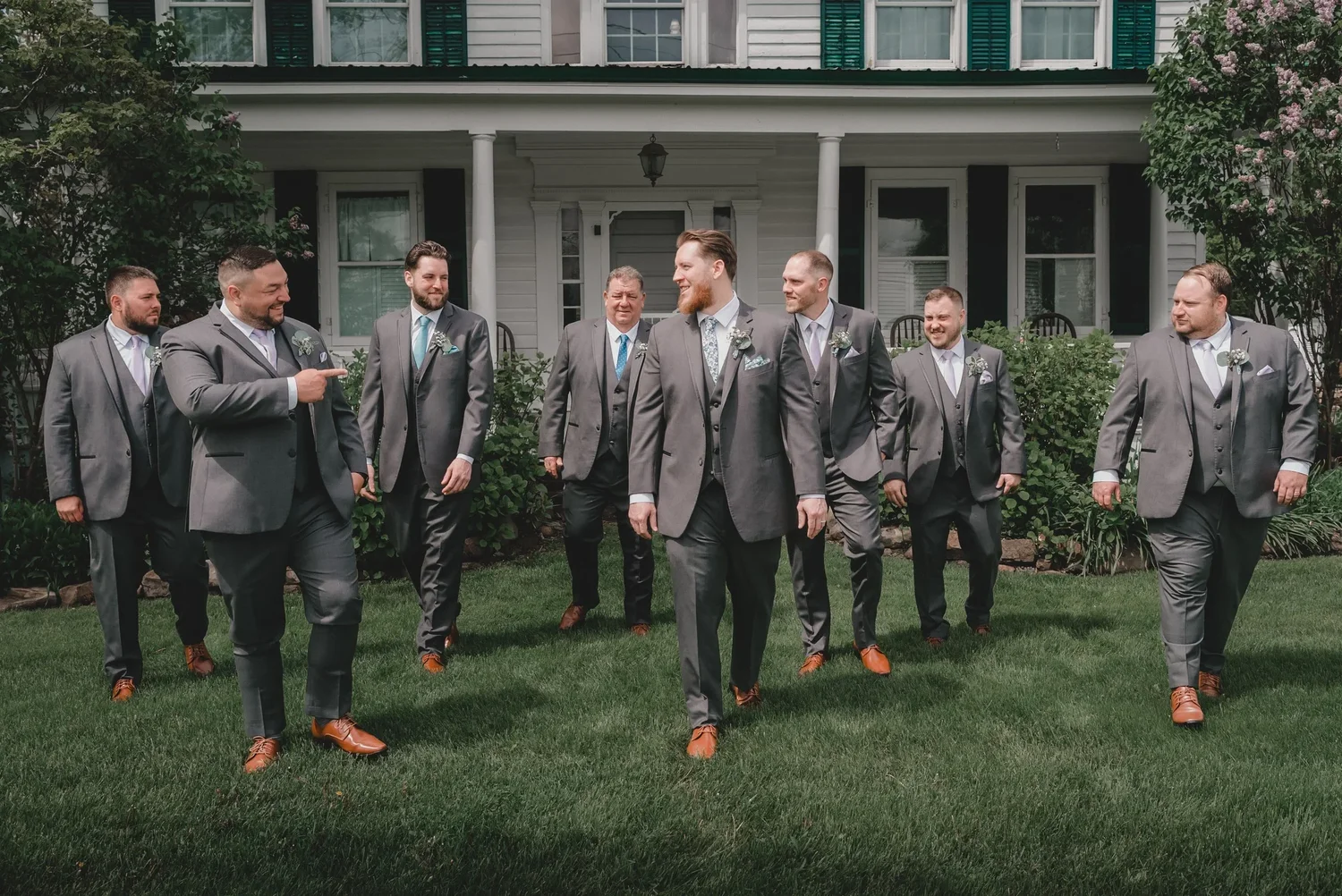 Christian and his groomsmen in grey suits walking across the lawn in front of the farmhouse, one of them laughing
