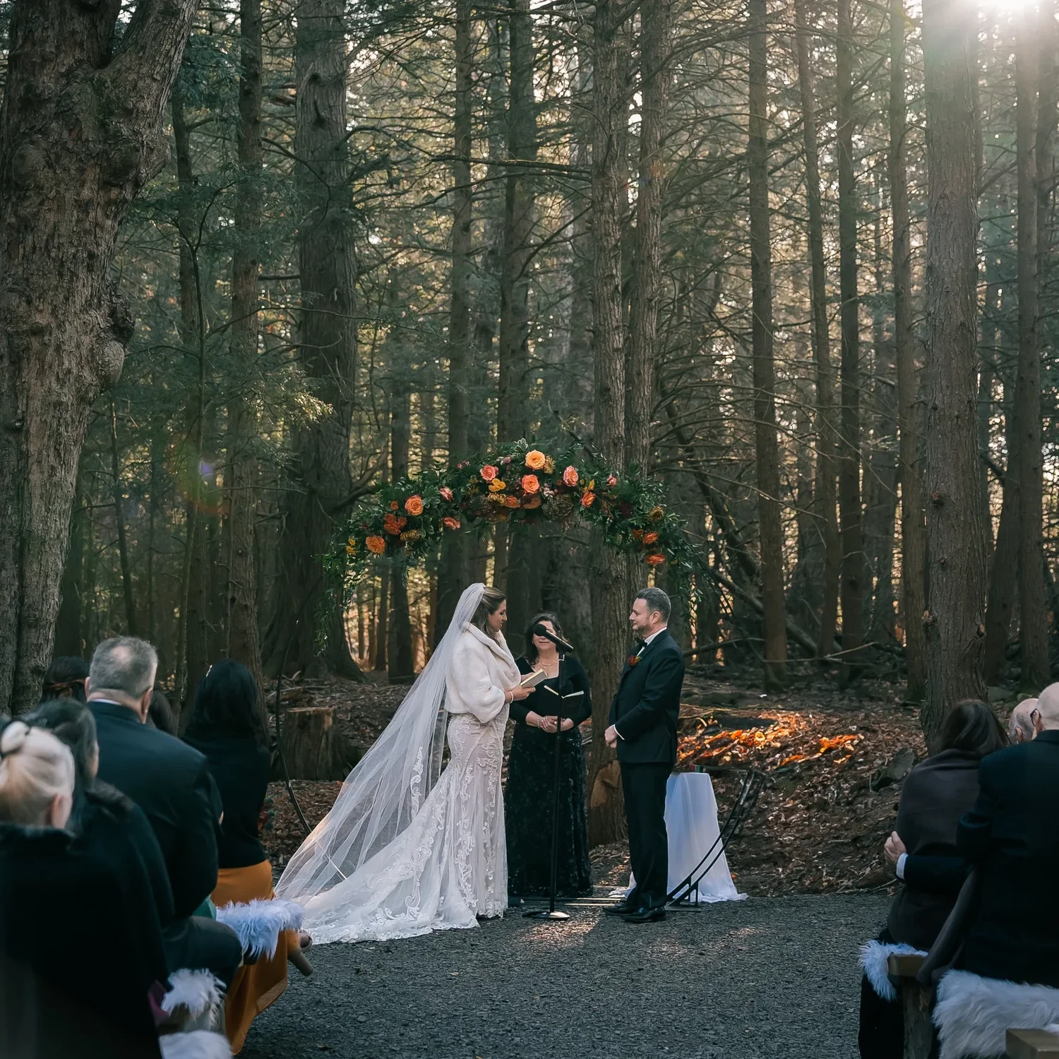 Charissa and Andrew under a floral arch in the Enchanted Forest, the officiant beside them and guests on wooden benches among the tall pines