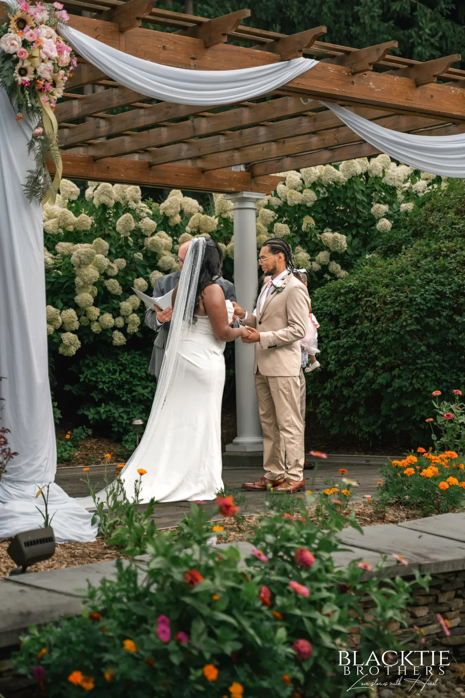 A bride and groom holding hands at their ceremony under a white-draped pergola at Traditions at the Glen, surrounded by white hydrangeas and summer flowers