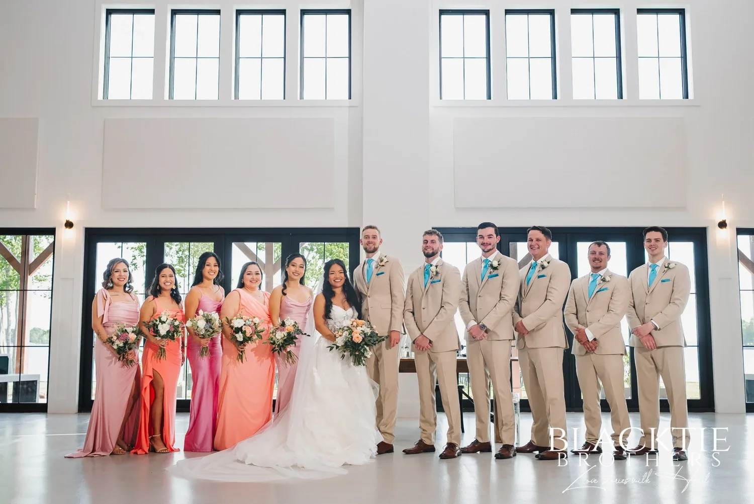 The full wedding party standing in a straight line in the main hall at Sally Port View, bridesmaids in shades of pink and coral holding bouquets and groomsmen in tan suits with teal ties, the tall black-framed glass doors and clerestory windows behind them