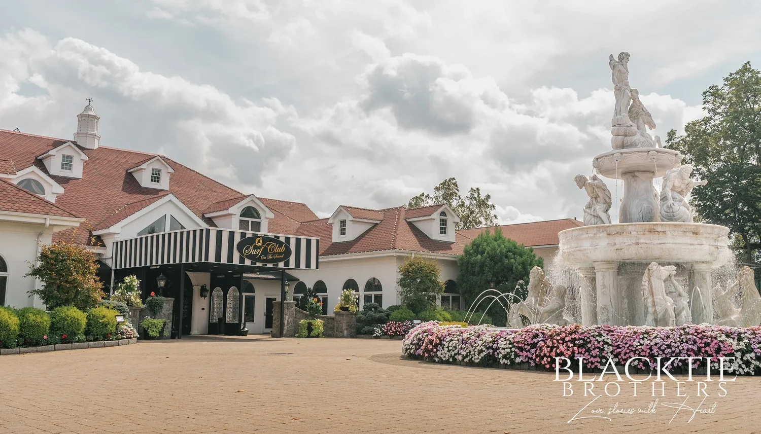 The front of The Surf Club on the Sound: a white mansion with a red roof and a striped awning over the entrance, a tiered marble fountain with statues and a bed of pink flowers in the circular drive
