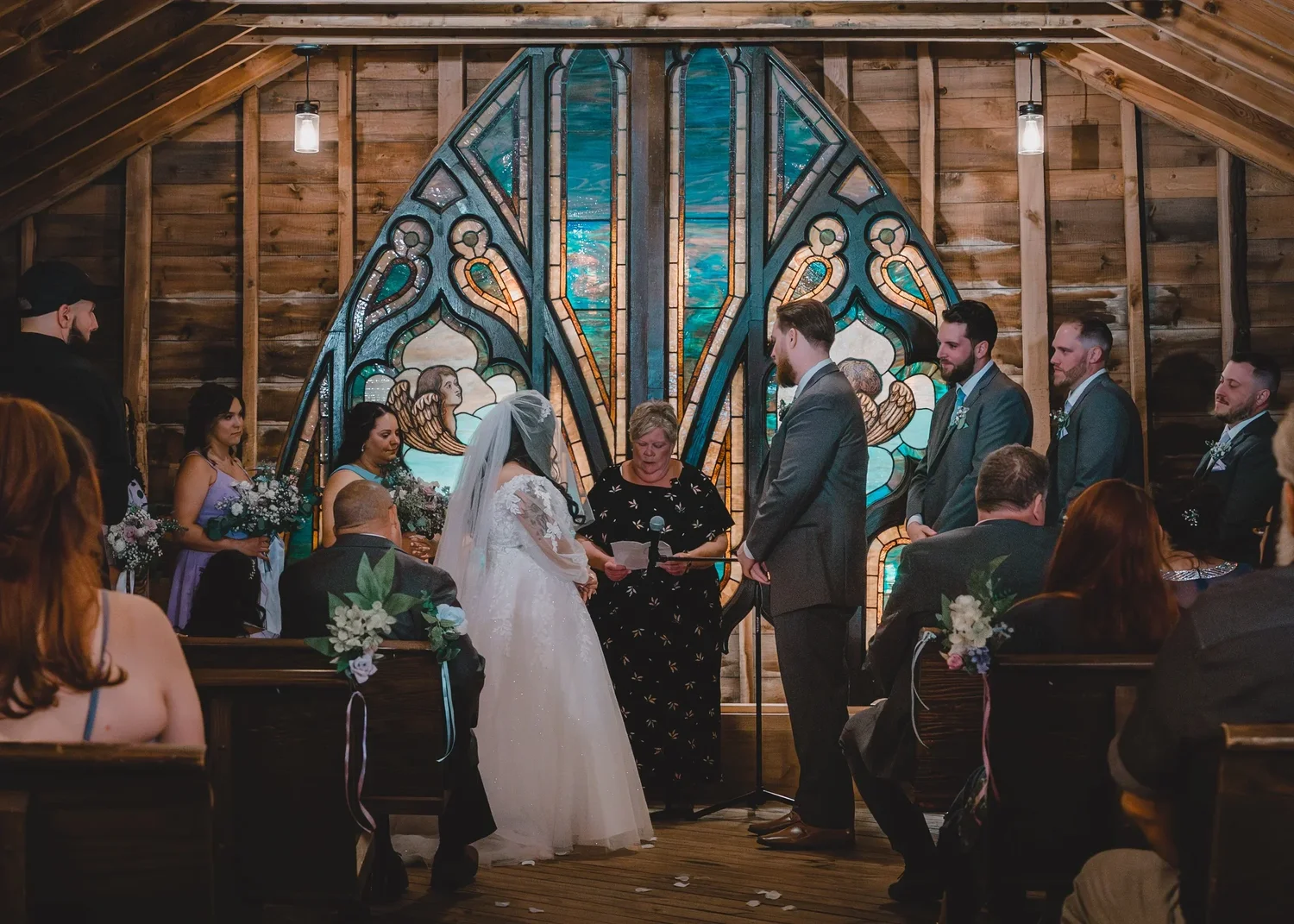 Jessica and Christian facing each other during their ceremony in the Loft Chapel, with the stained glass window behind them and guests in the pews