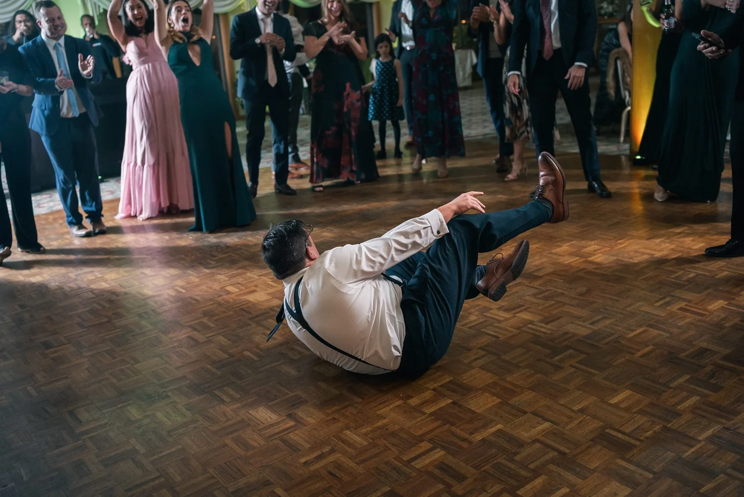 A guest in suspenders spinning on his back in the middle of the dance floor while everyone around him cheers