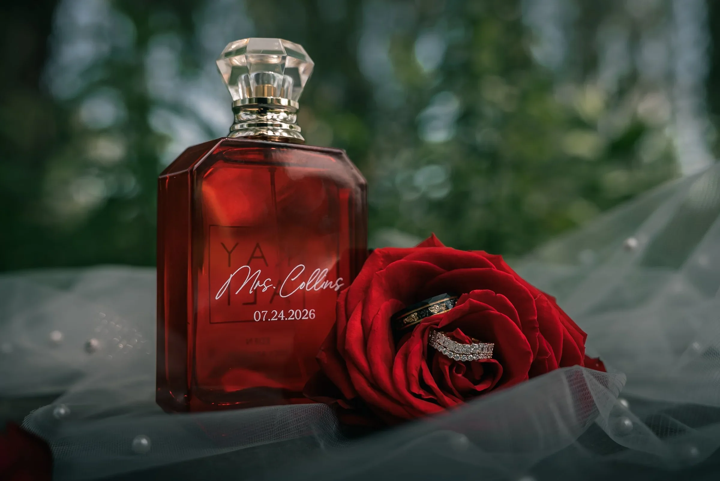 A red perfume bottle engraved with Vanessa’s new name and wedding date, beside a red rose holding both wedding rings