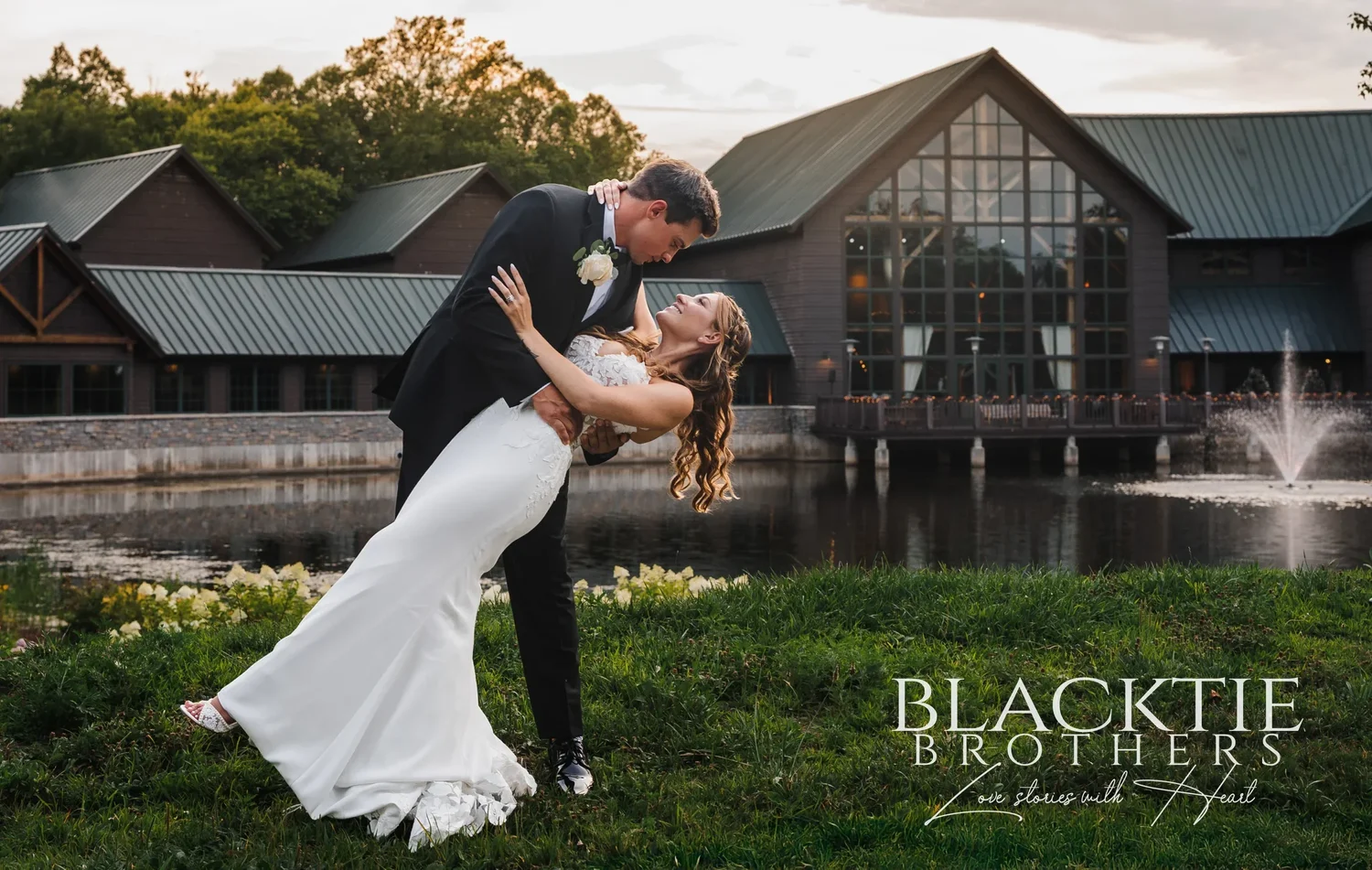 A couple on the far bank of the pond at Allyn Lodge in Skaneateles Falls, New York, with the timber lodge and its fountain behind them at sunset.