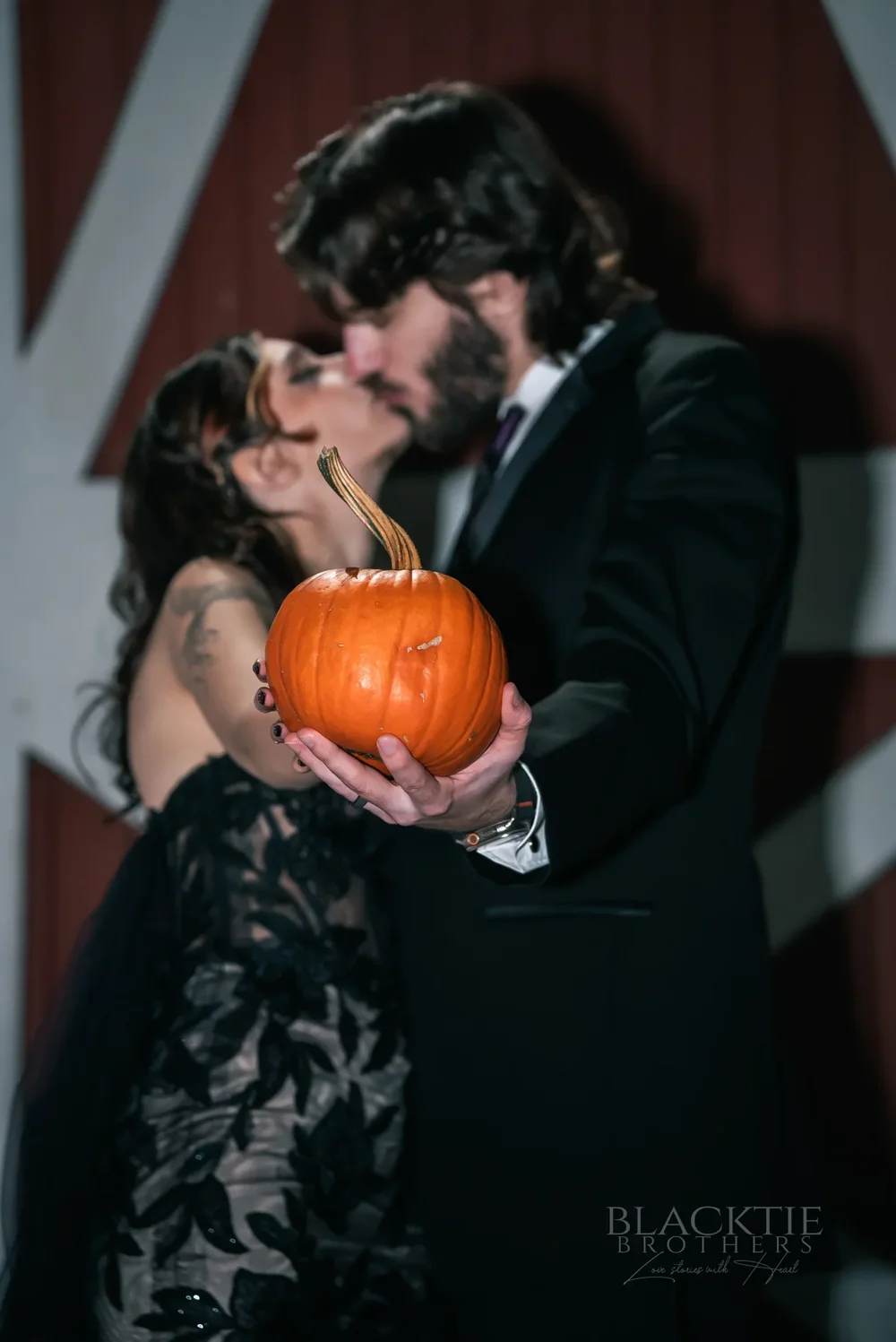 Maddy and Dan kissing behind a pumpkin in front of a red barn door at their Halloween wedding