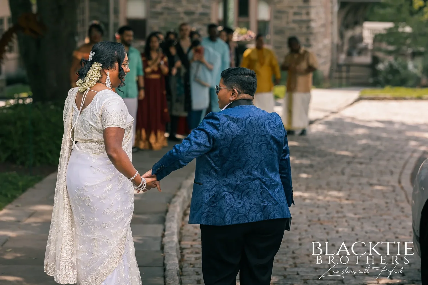 Hima and Alina walking hand in hand up a cobbled drive toward a crowd of guests in bright clothes outside a stone building, one guest clapping
