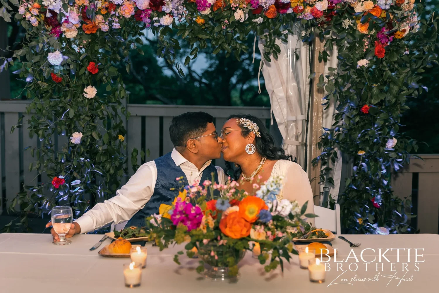 Hima and Alina kissing at their sweetheart table under the flower arch, candles and flowers on the table in front of them